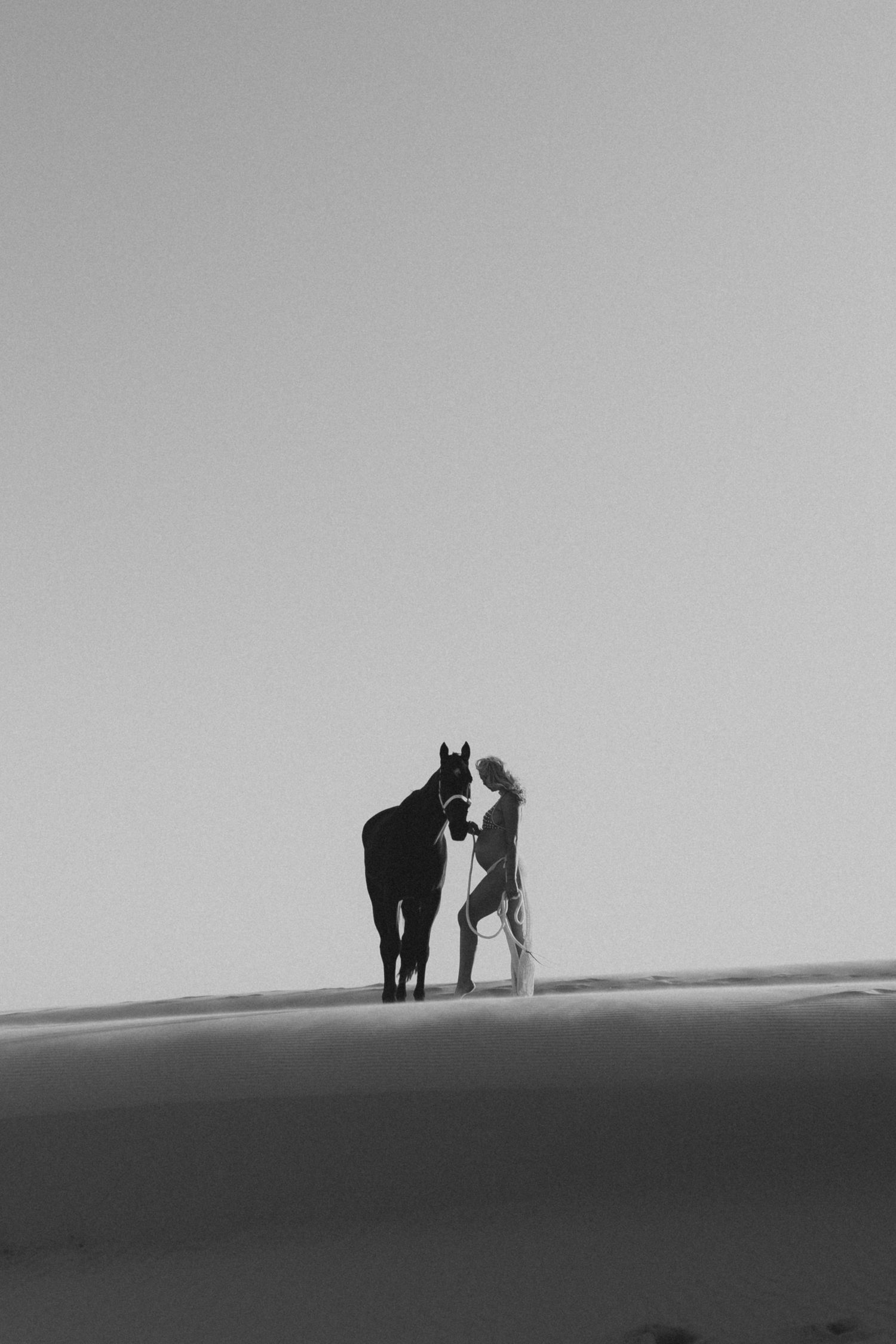 Black and white photo of a woman standing with a horse on desert sand dunes.