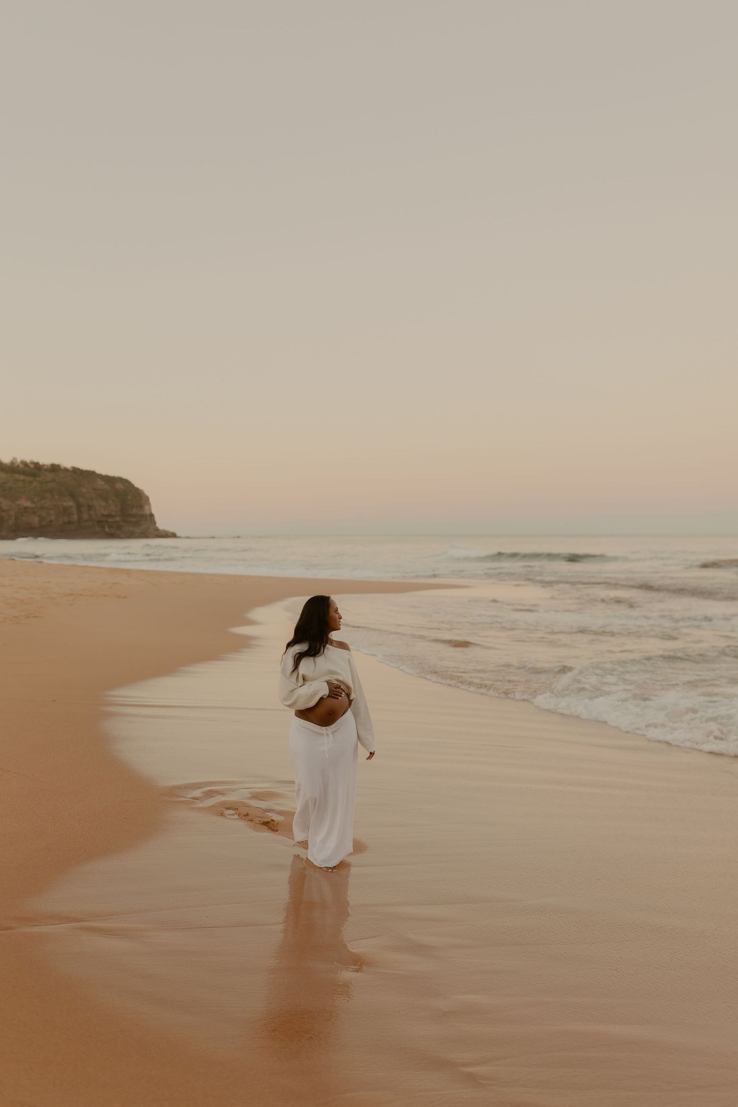 Pregnant woman in white dress walks along sandy beach shore at golden sunset.