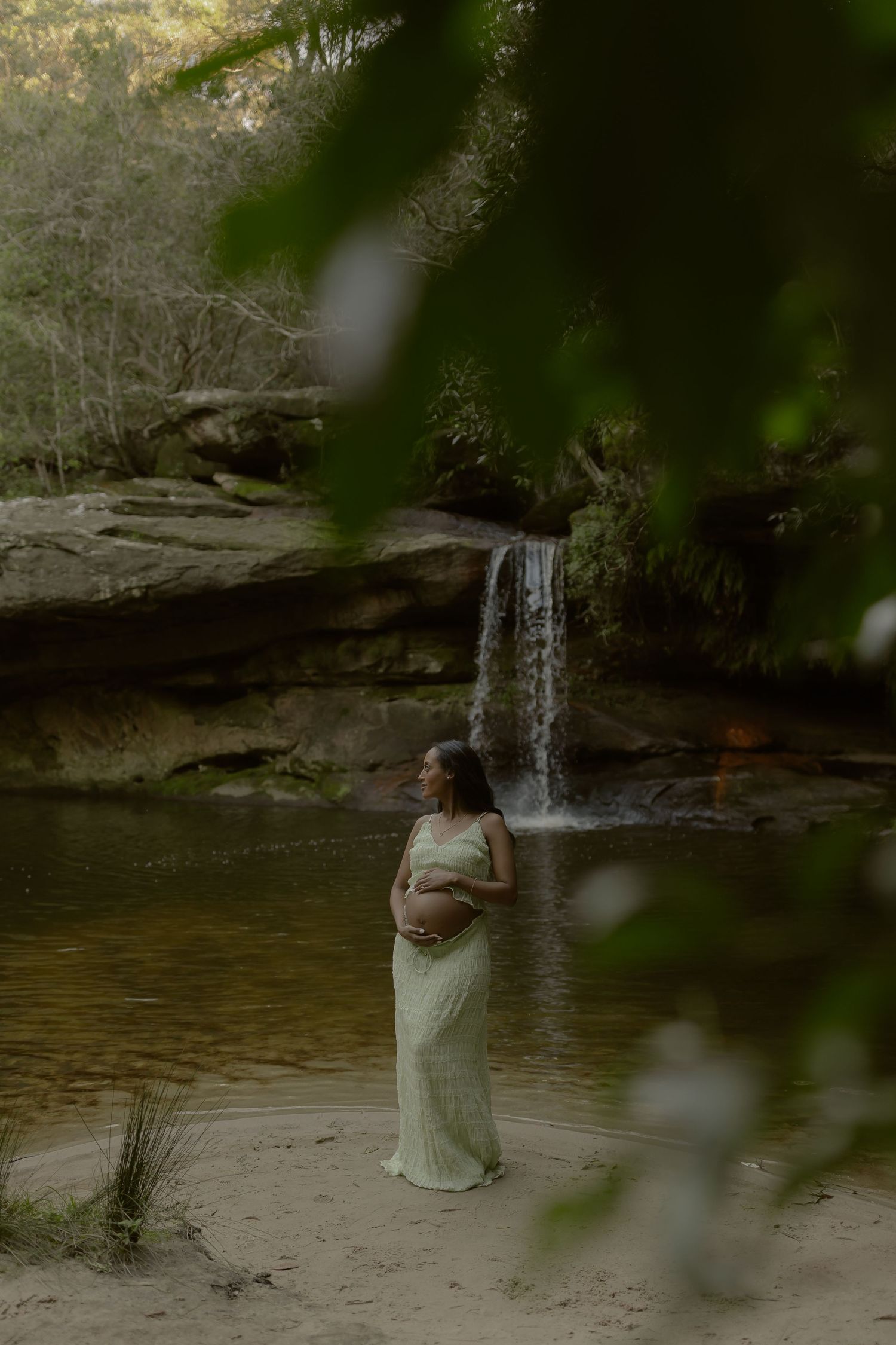 Pregnant woman in green dress cradling belly by a serene waterfall in a lush forest setting.