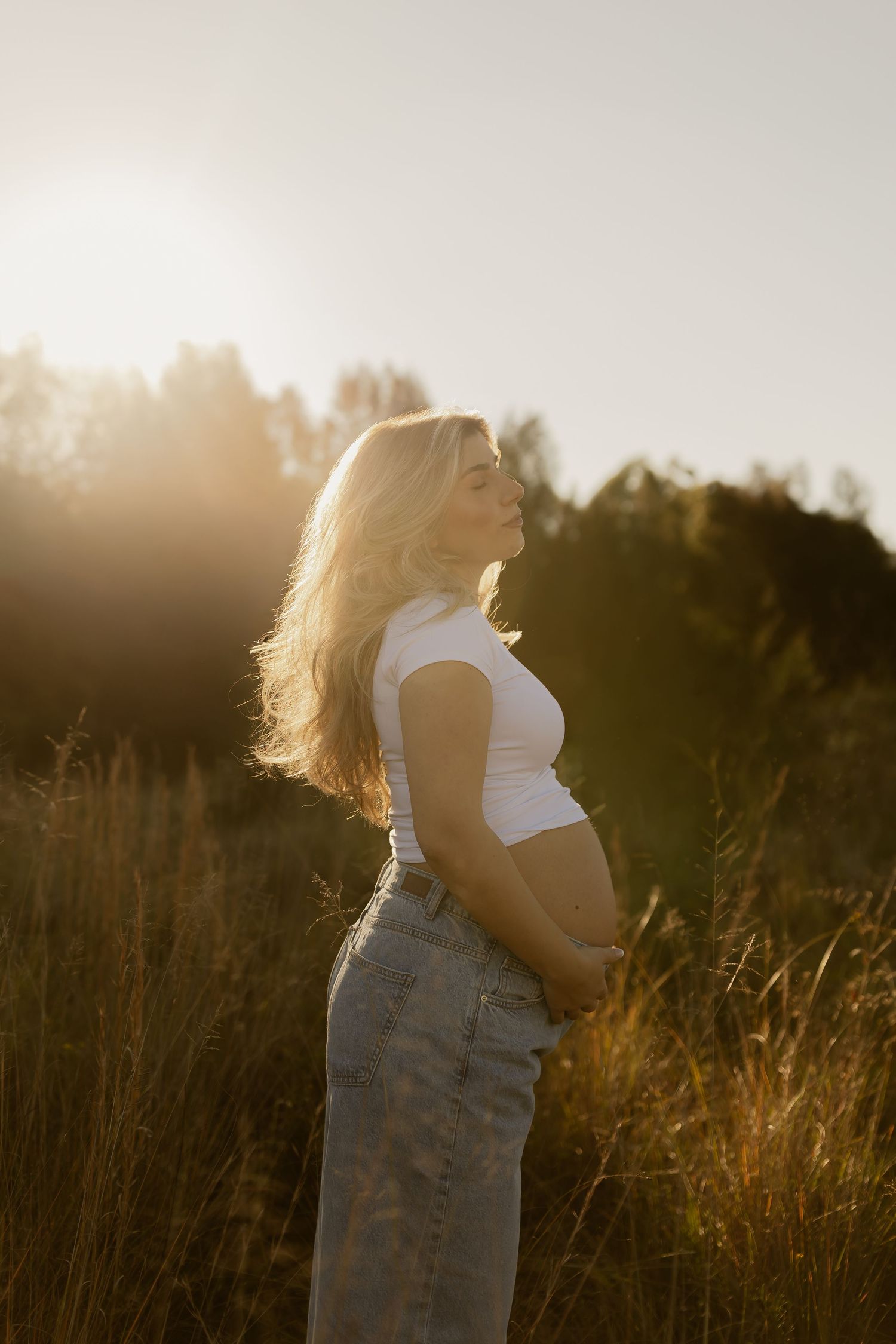 Pregnant woman in white crop top and jeans stands in golden sunlit field, cradling her baby bump.