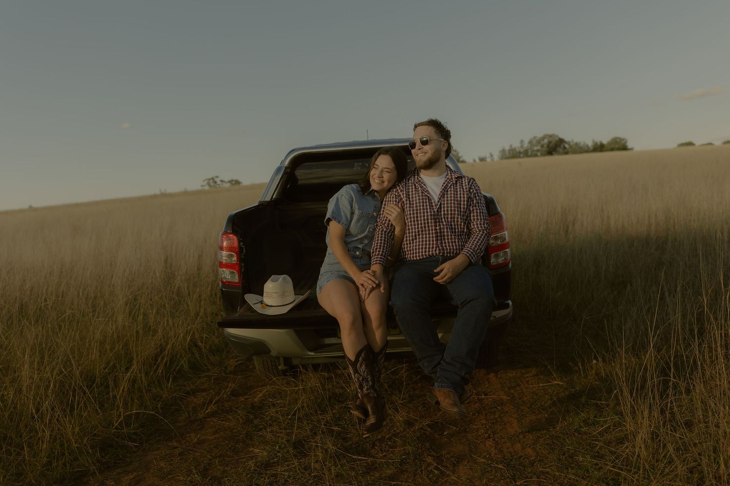 Couple sitting on truck tailgate in golden field at sunset, woman in boots and man in sunglasses, rural countryside.
