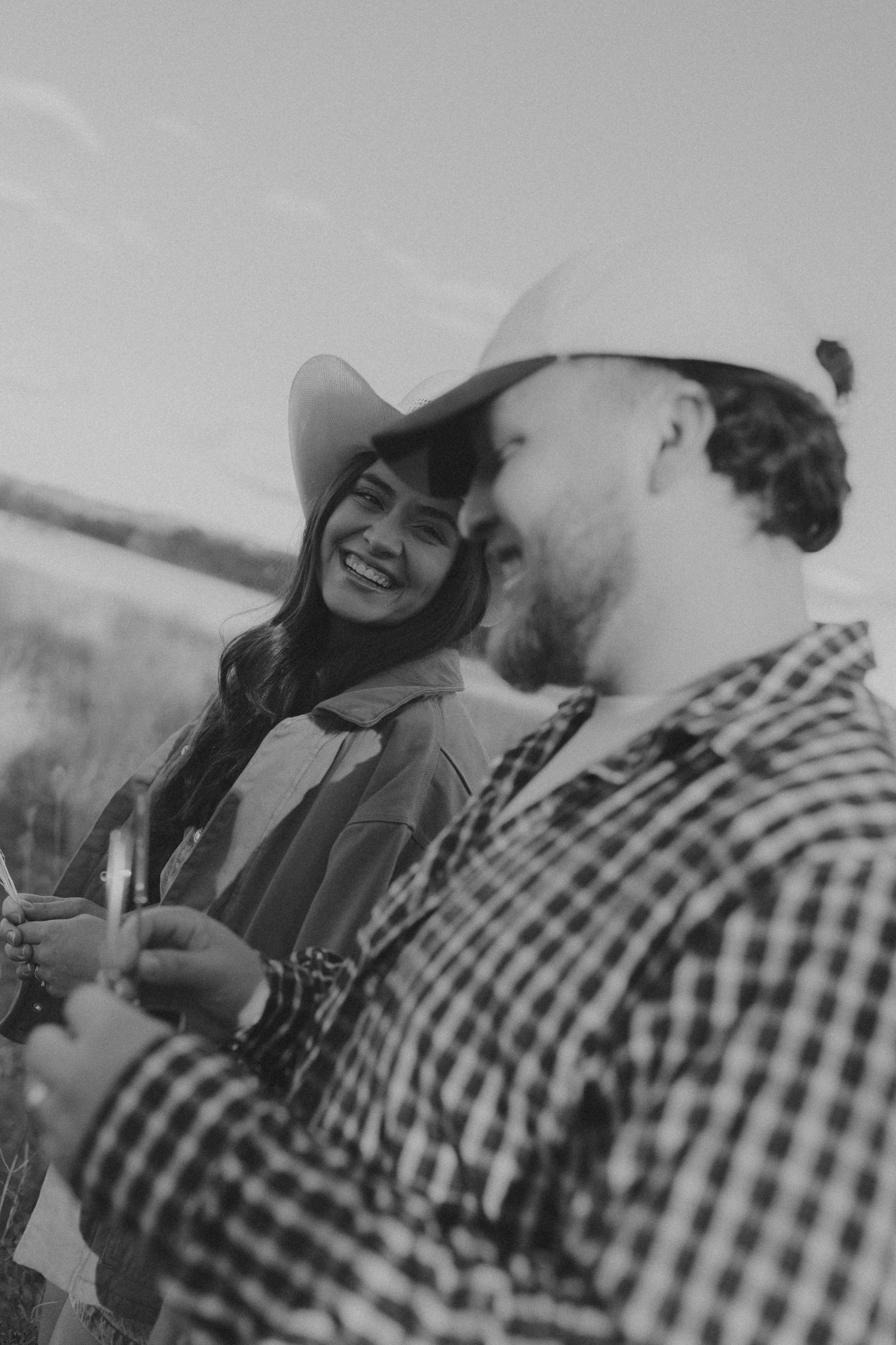Black and white photo of a smiling couple wearing cowboy hats outdoors, woman gazing at man in plaid shirt.