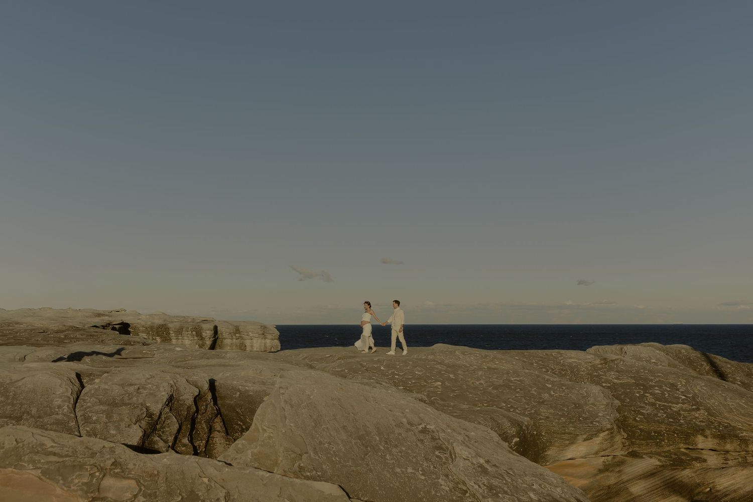 Two people in white stand on rocky coastal cliffs overlooking a calm blue sea under a clear sky.