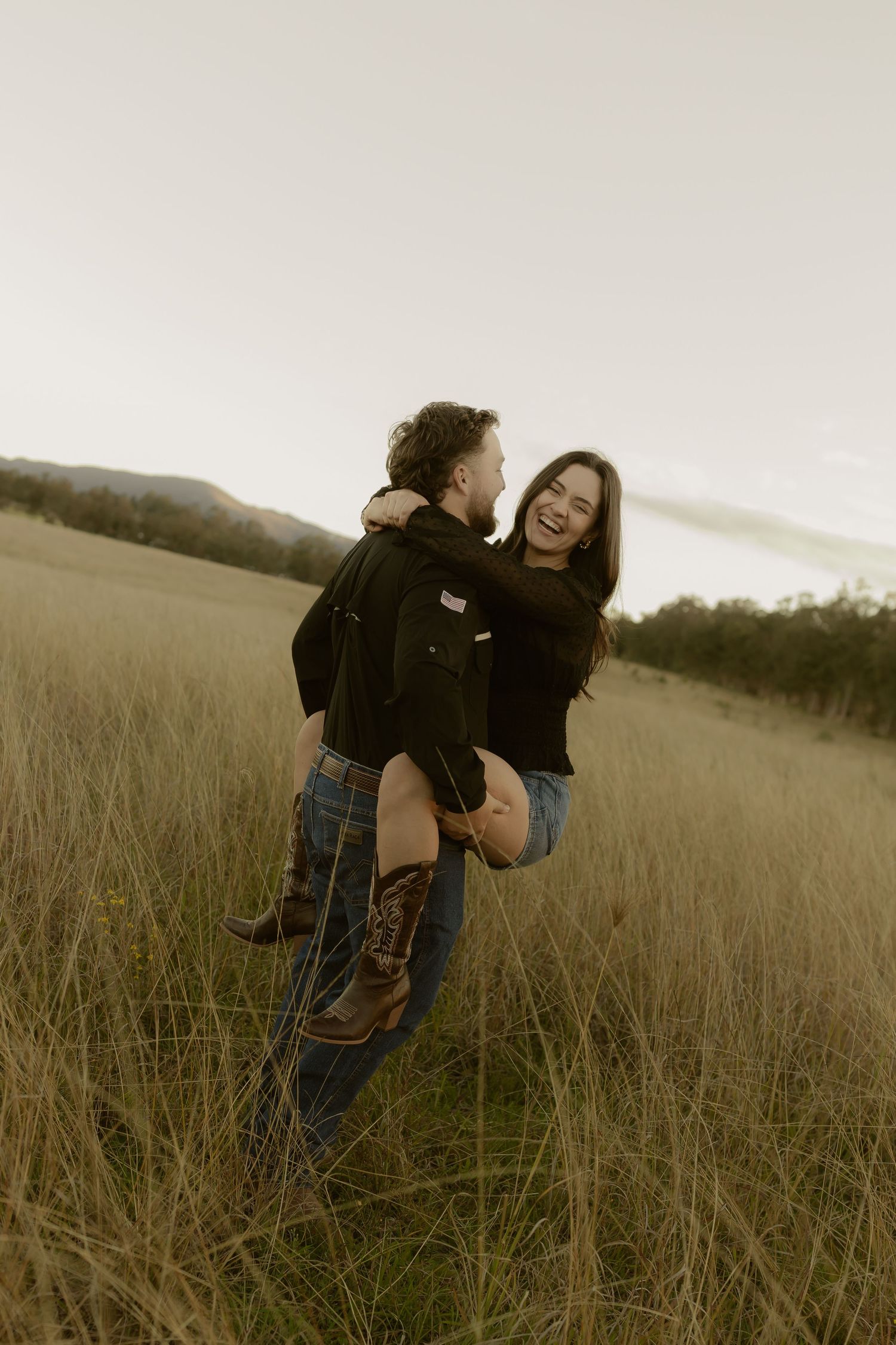 A joyful couple embraces in a golden field, man lifting woman as she laughs, with rolling hills at dusk.