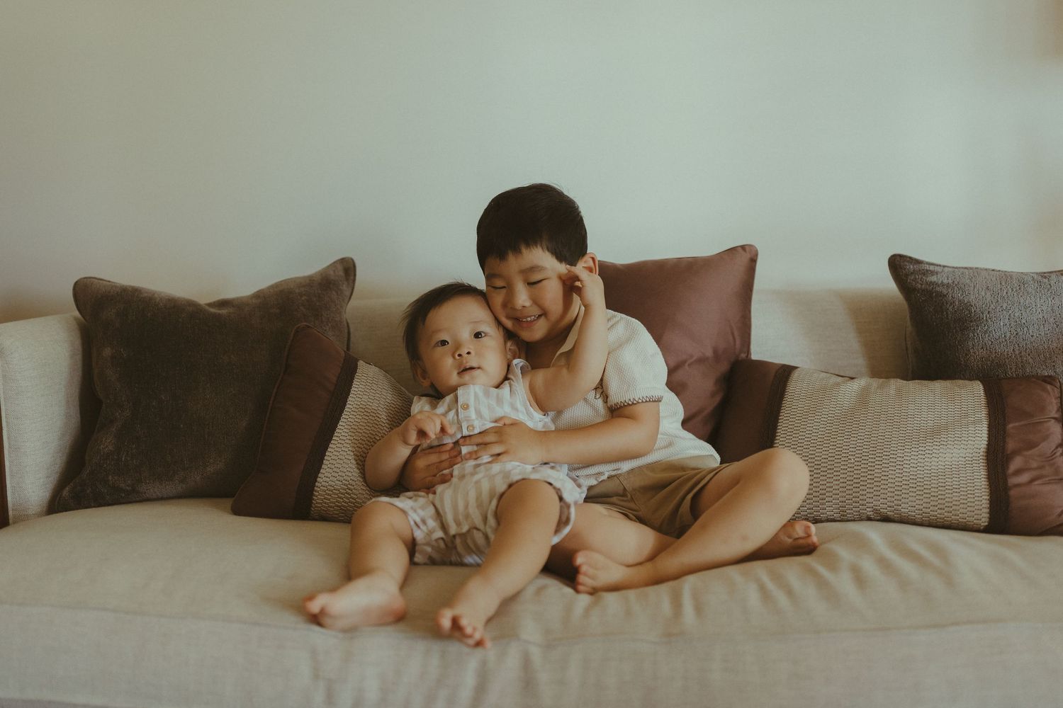 A parent and child share a joyful moment while sitting together on a beige couch with brown throw pillows.