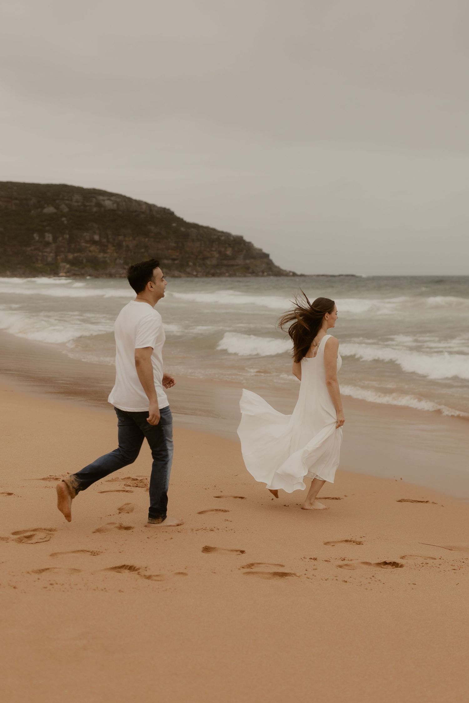 A couple in white clothing chases each other playfully along a sandy beach with cliffs in the background.