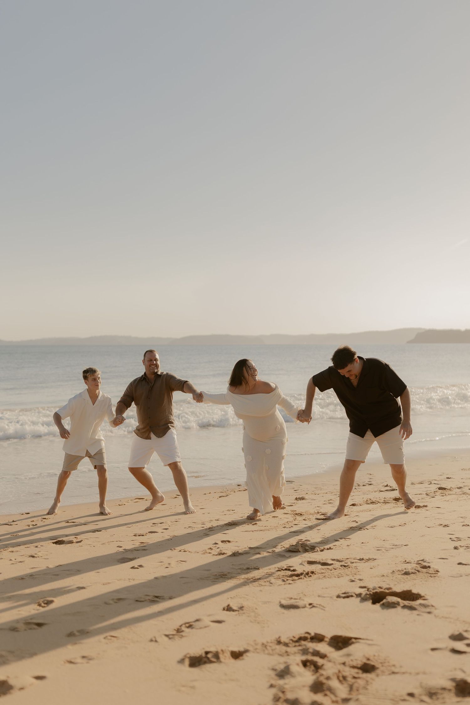 A group in white and black outfits playfully holding hands while dancing on a sunny beach at sunset.