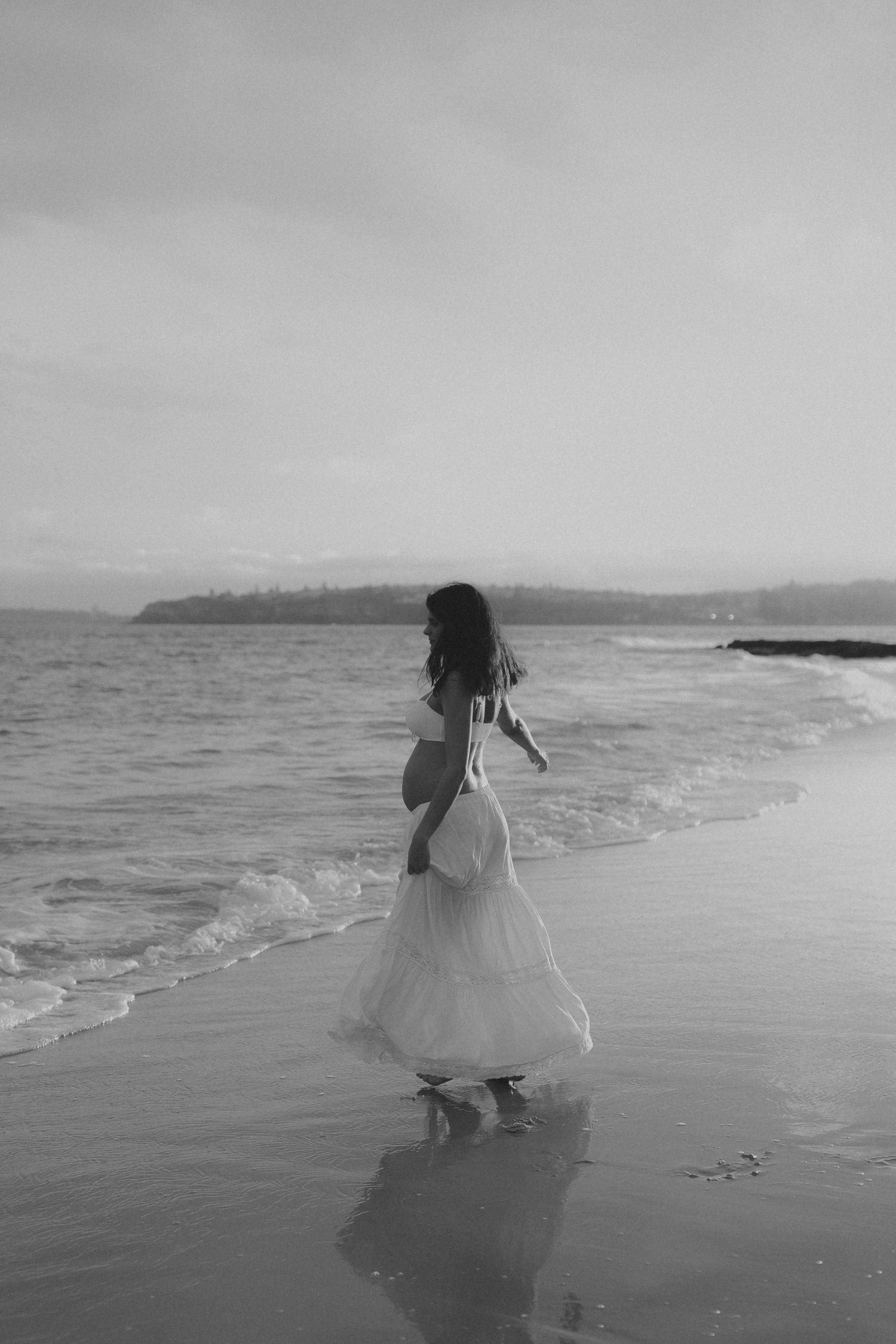 A figure in a flowing white dress dances along the shoreline in black and white.