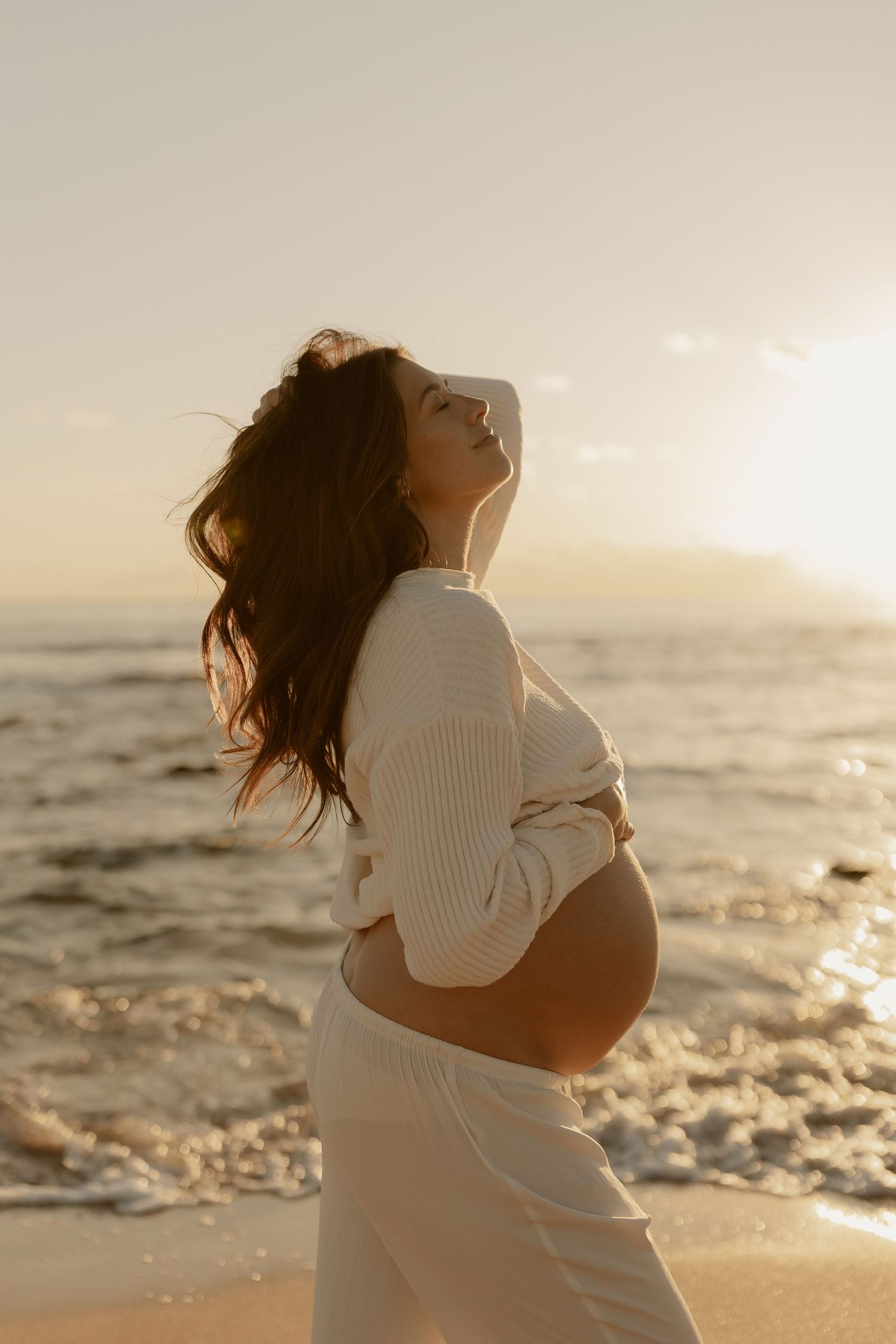 Woman in white sweater dancing joyfully on beach at golden hour with ocean waves in background.