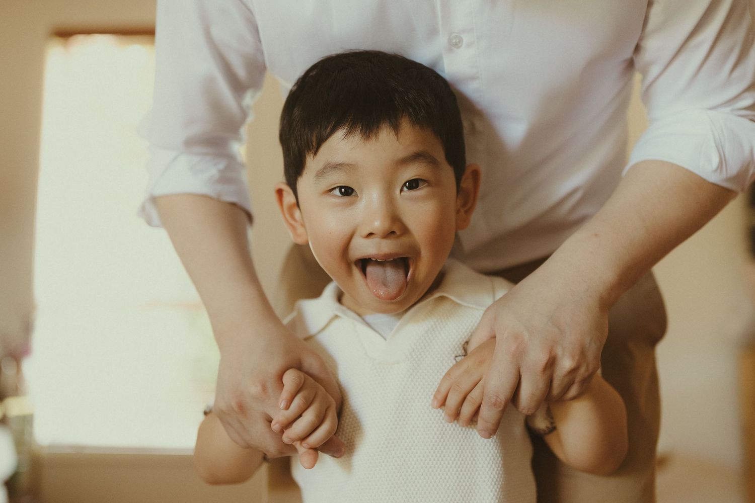 A young child in a white shirt shows excitement while adult hands rest on their shoulders in a warm-toned indoor setting.
