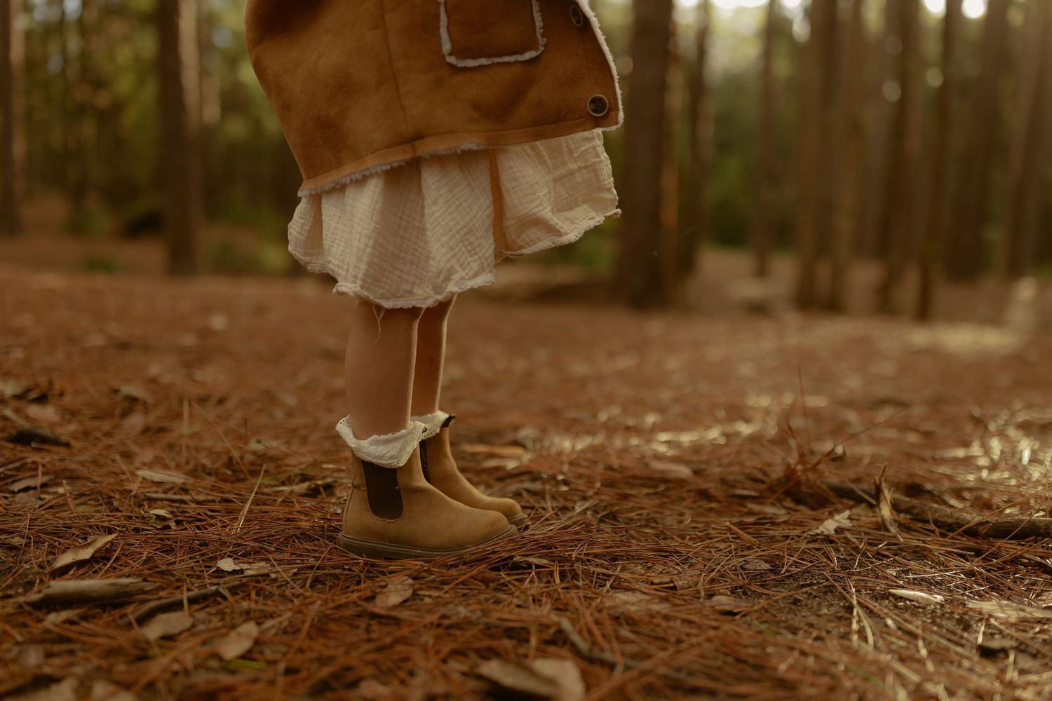 Close-up of young child wearing white dress and tan boots standing on forest floor.