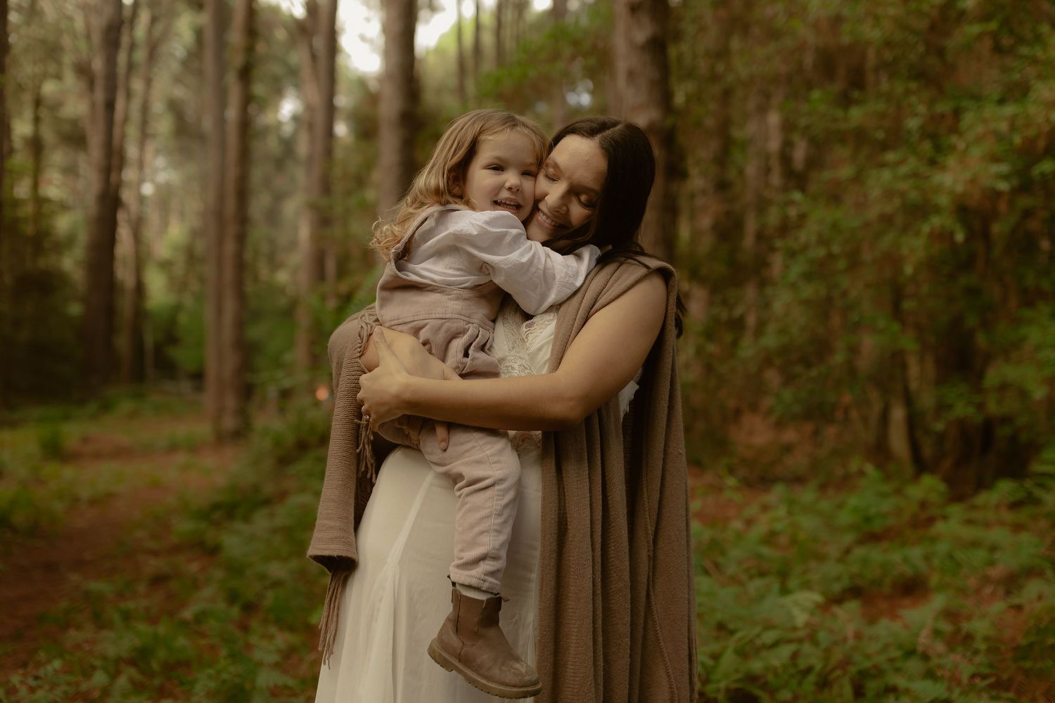 A mother tenderly embraces her toddler in a sunlit forest, both wearing soft neutral tones.