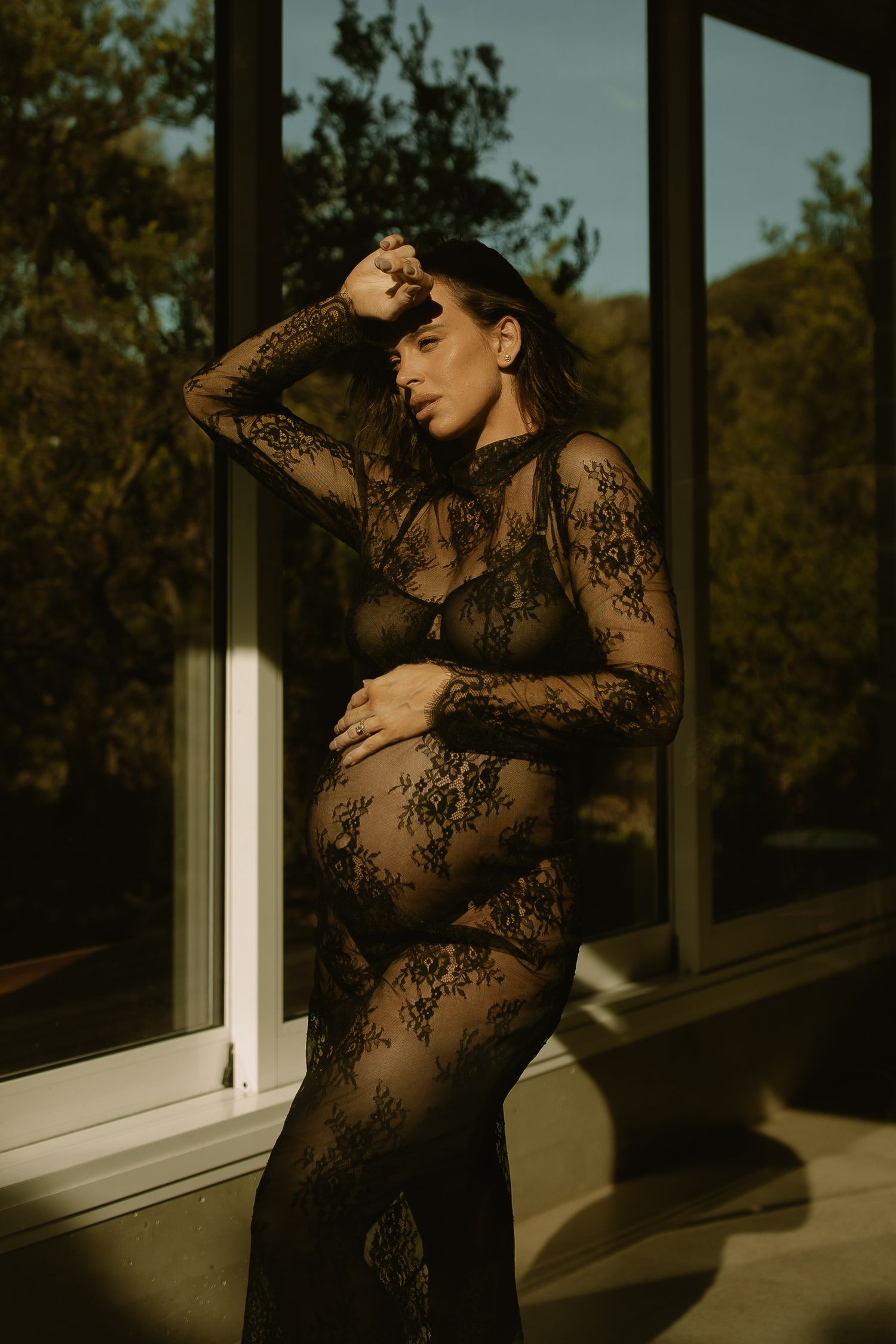A silhouetted figure in a sheer lace dress poses by a window in warm sunset light creating a dramatic maternity portrait.