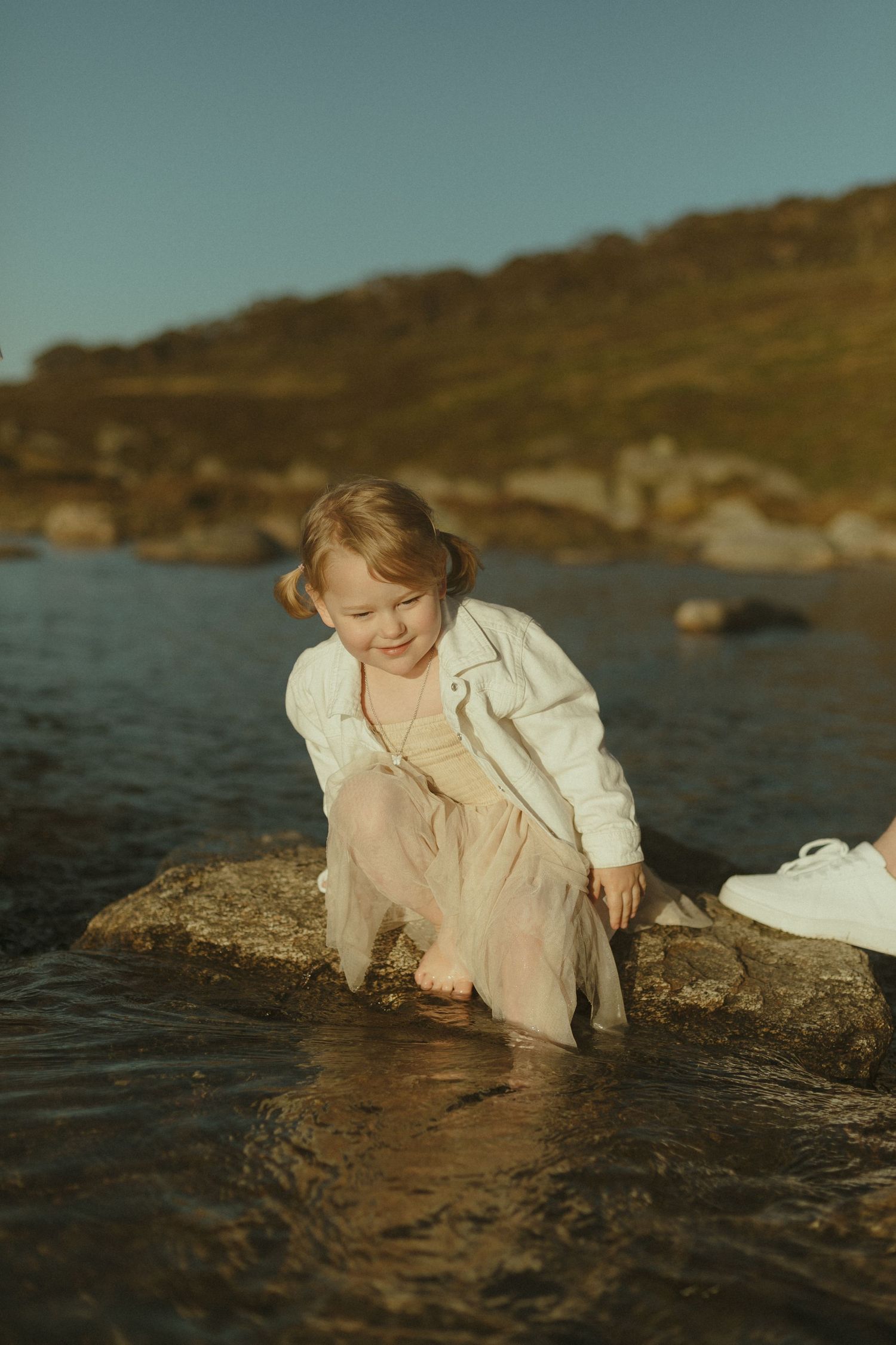 A child in a white jacket and light dress plays at the water's edge on a rocky coastline at sunset.
