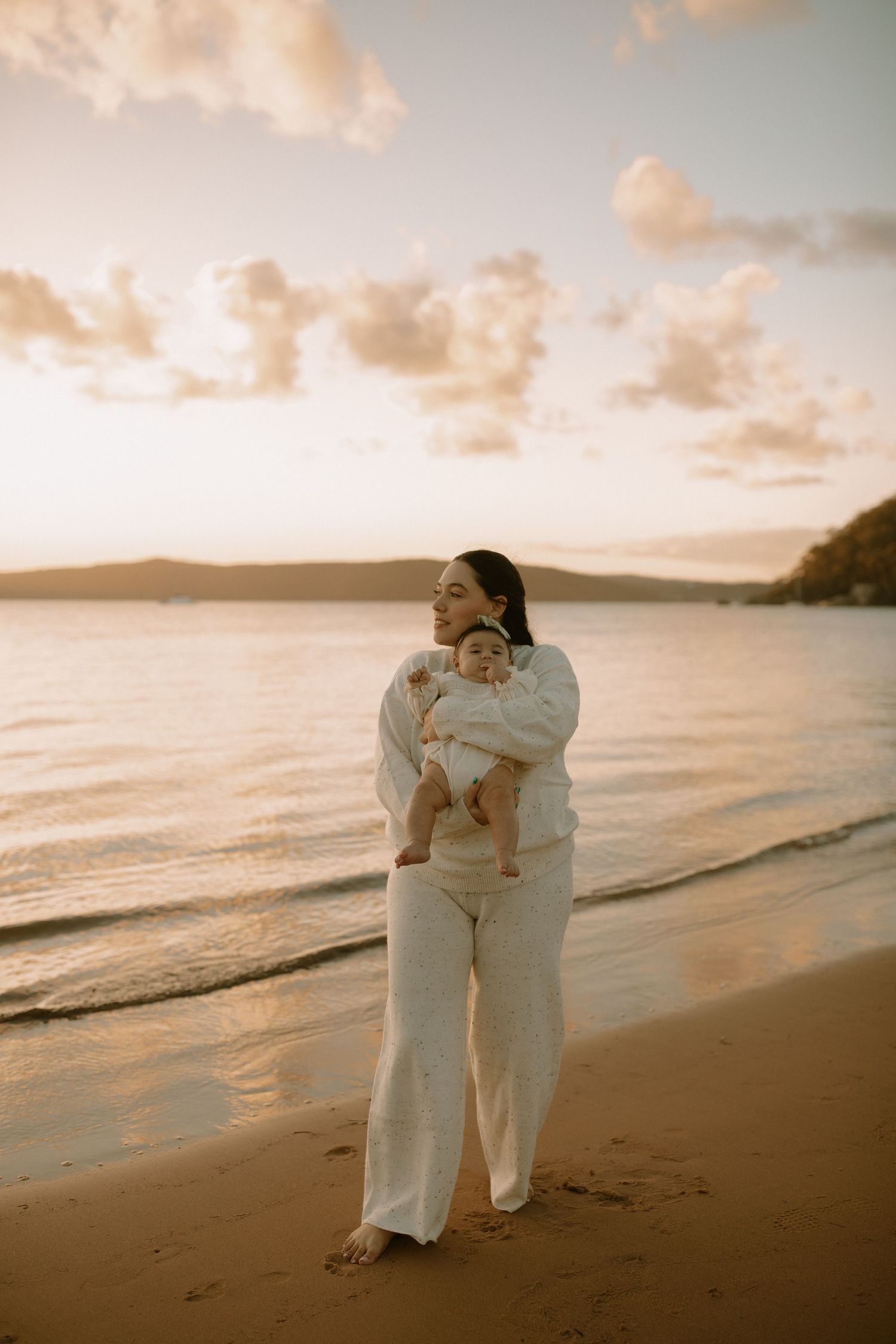 A person in white clothing stands on a sandy beach at sunset with calm waters and soft clouds in the background.
