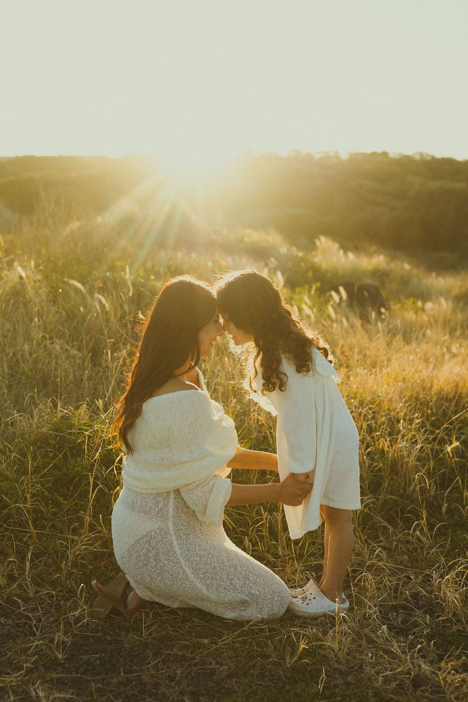 Two figures in white dresses sit together in a sunlit field of tall grass during golden hour.