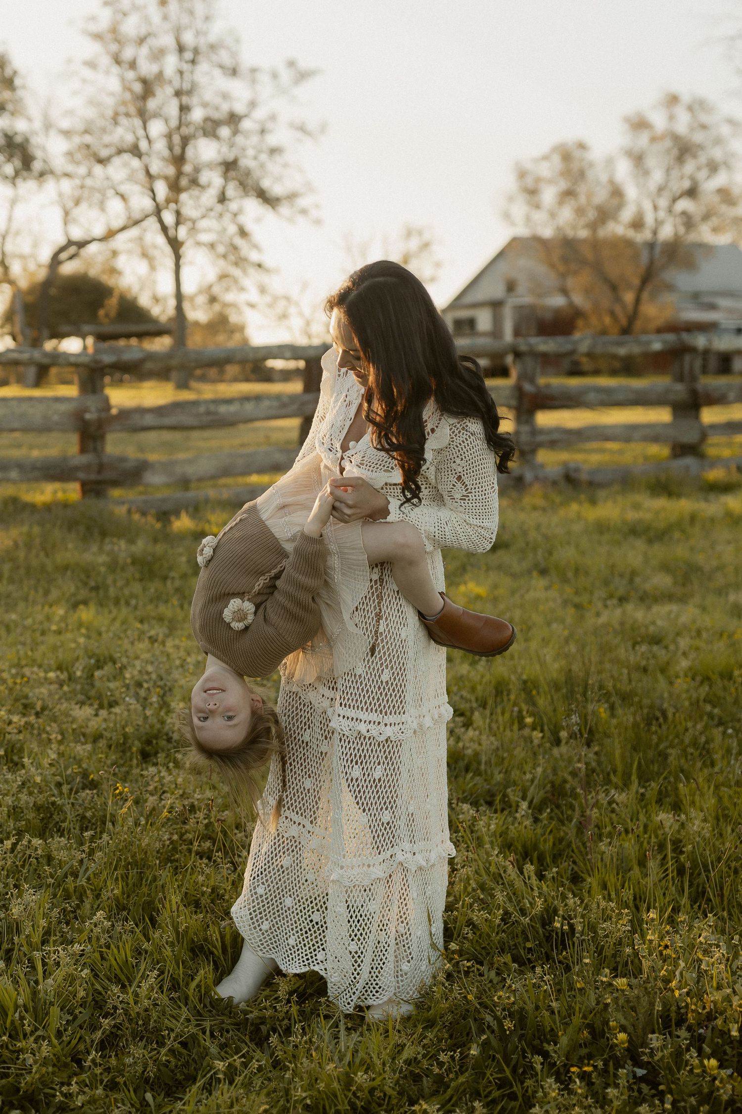 Woman in white lace dress dancing playfully in a golden field at sunset near a farmhouse.