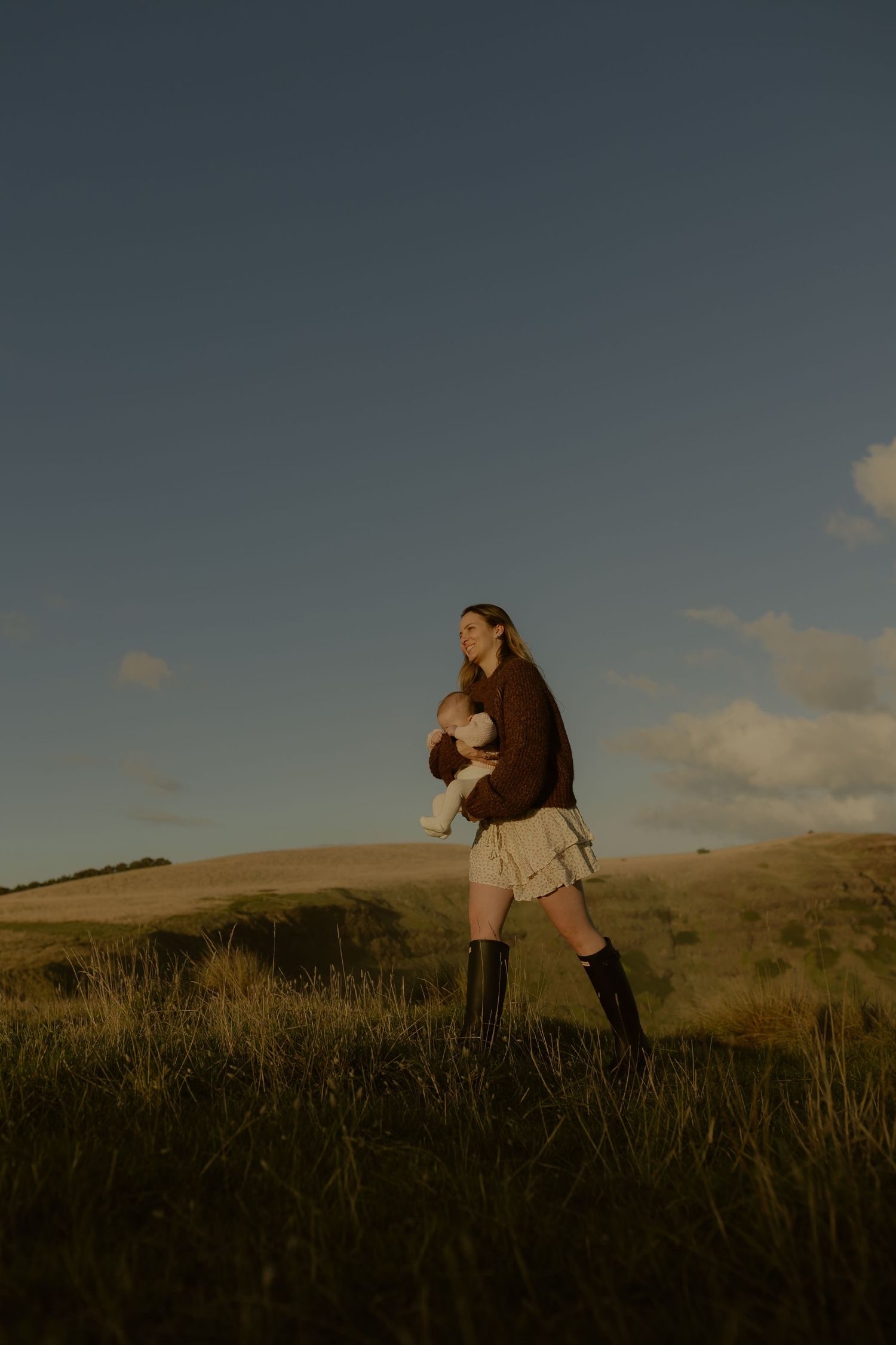 Woman in brown sweater and skirt holding a baby while standing in a golden grassy field at dusk.