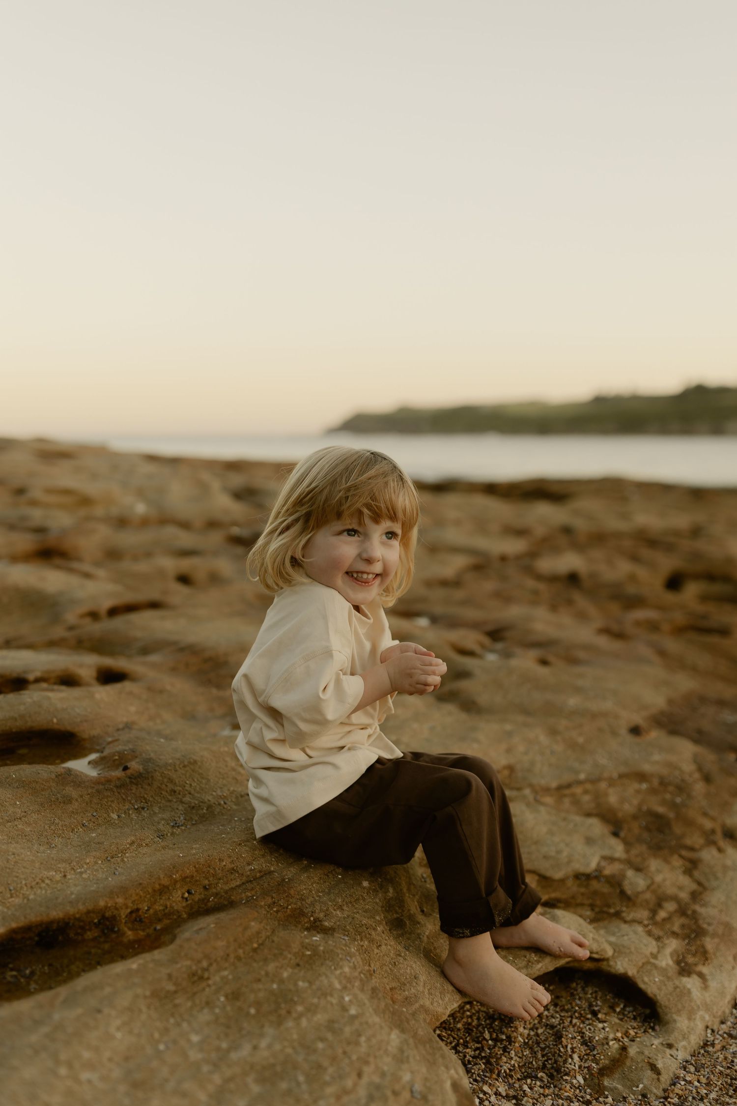 A smiling young child with blonde hair sits barefoot on rocky coastal terrain wearing a cream top and dark pants.