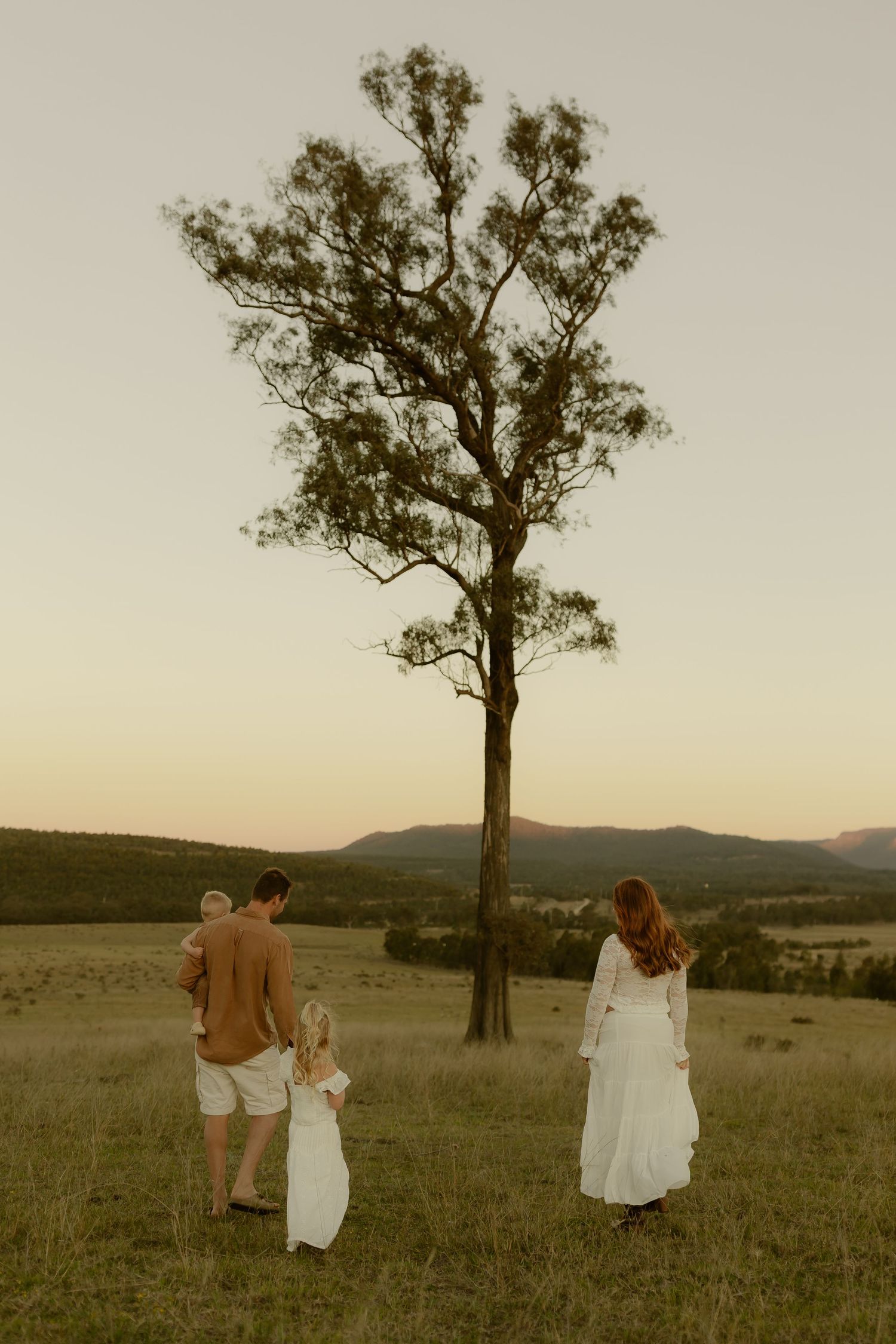 Family of four standing in a golden field at sunset near a tall tree with mountain views.