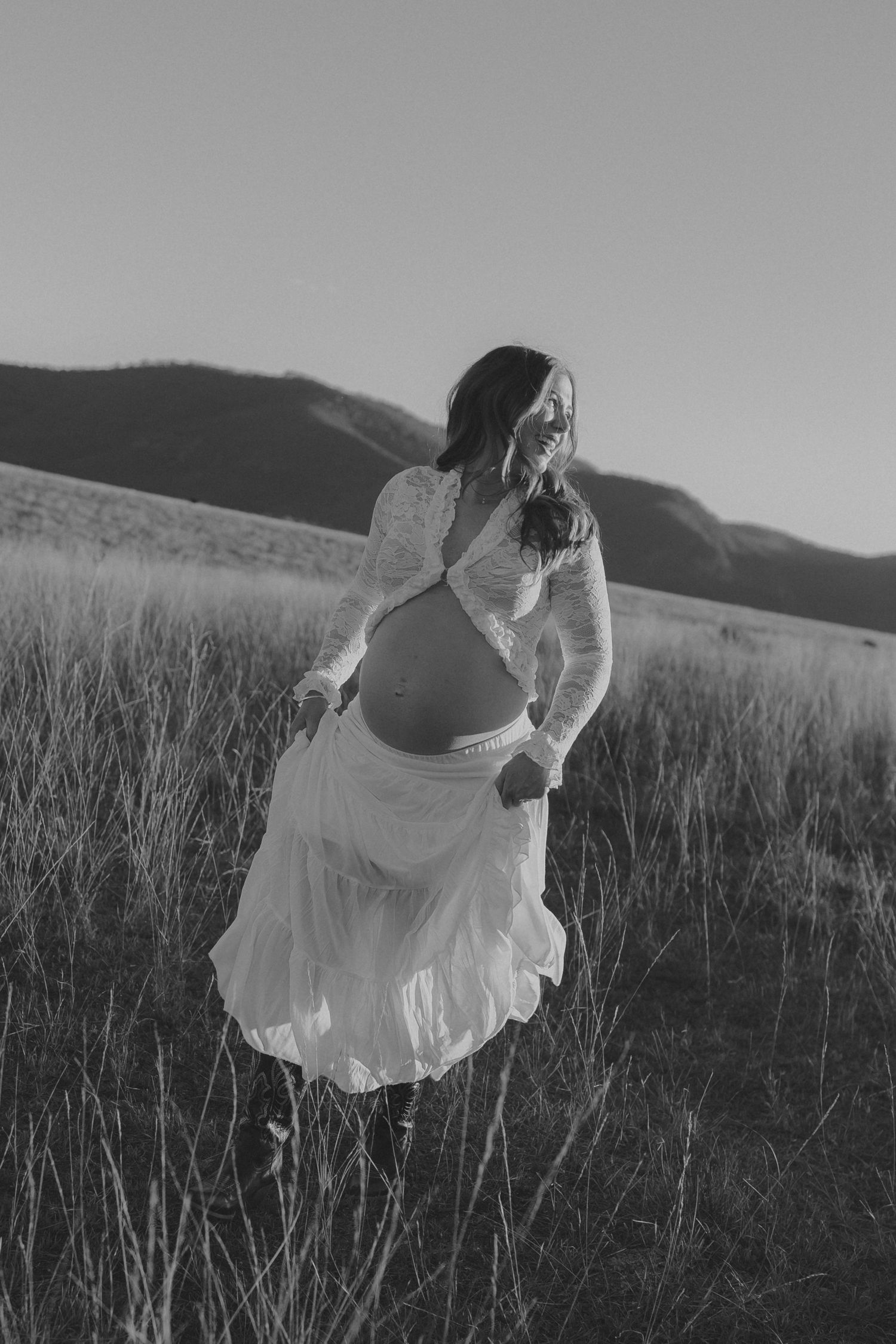 A pregnant woman in a white dress walks through a field, smiling, in a black and white photo.