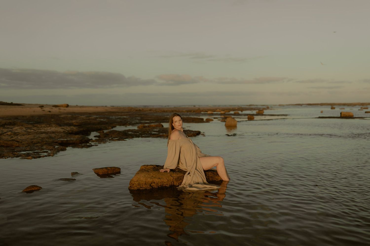 A dreamy coastal scene with someone perched on a rock surrounded by shallow tidal waters at dusk.