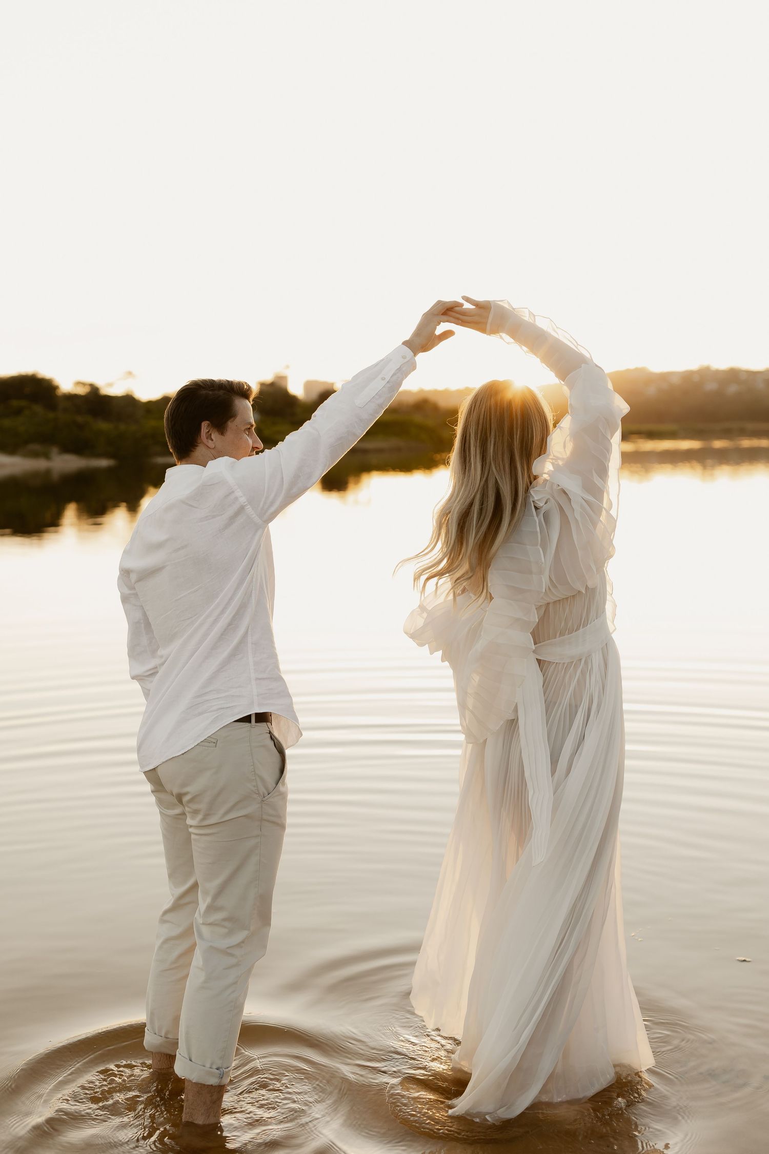 Couple in white clothing dancing together in shallow water during sunset.