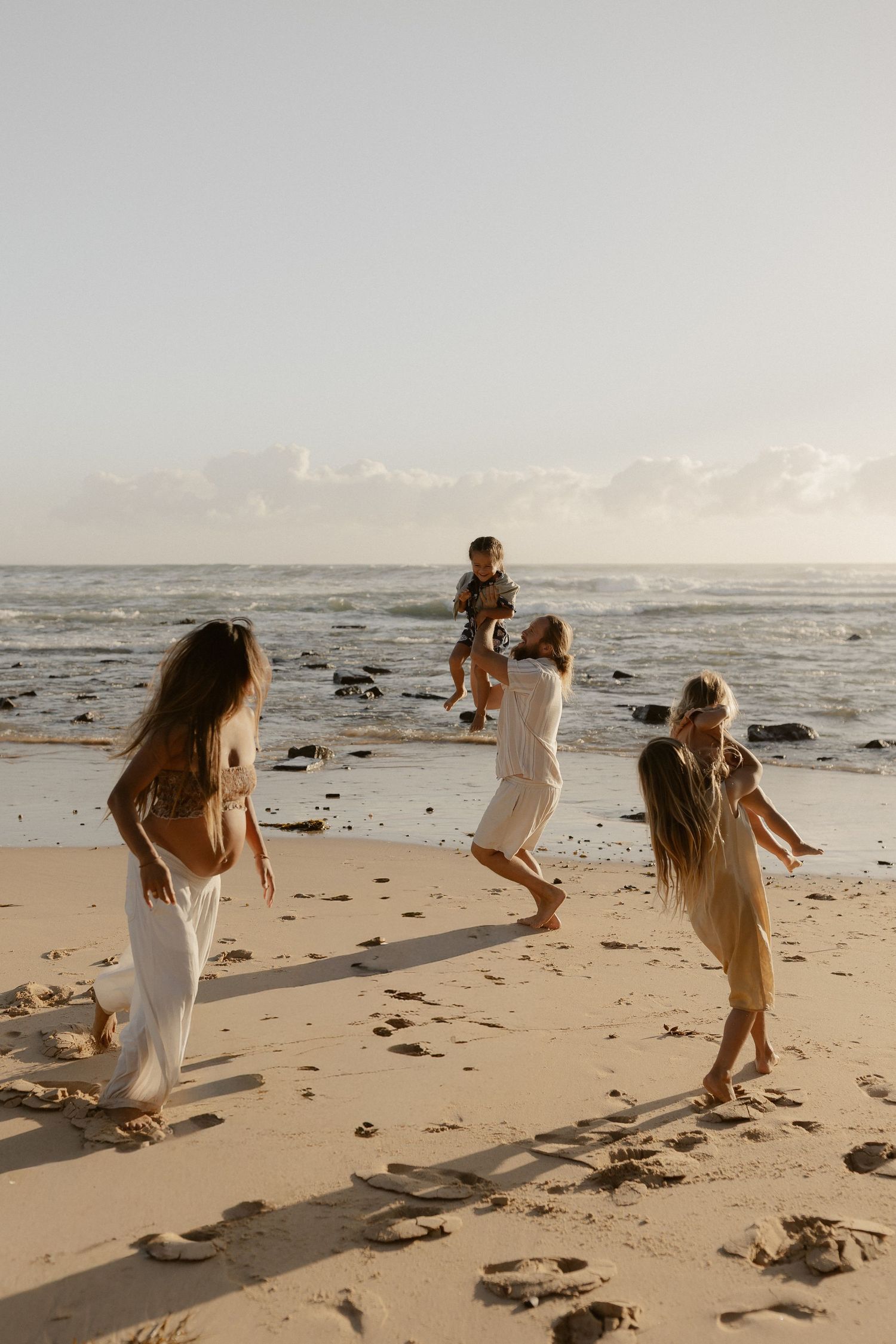 Group in white dresses dances and plays together on a sandy beach at golden hour.