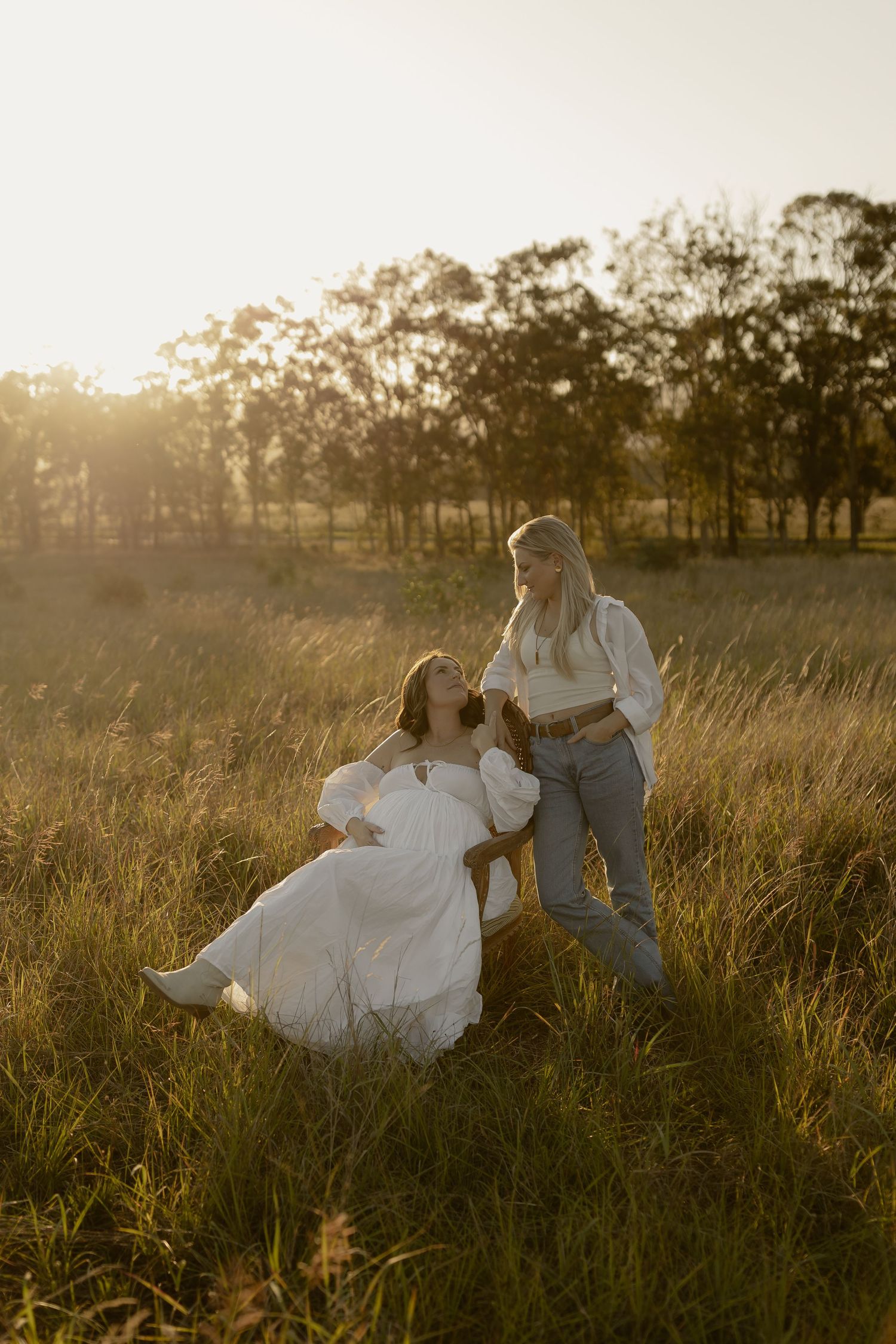 A romantic couple sits in a sunlit meadow during golden hour, surrounded by tall grass and trees.