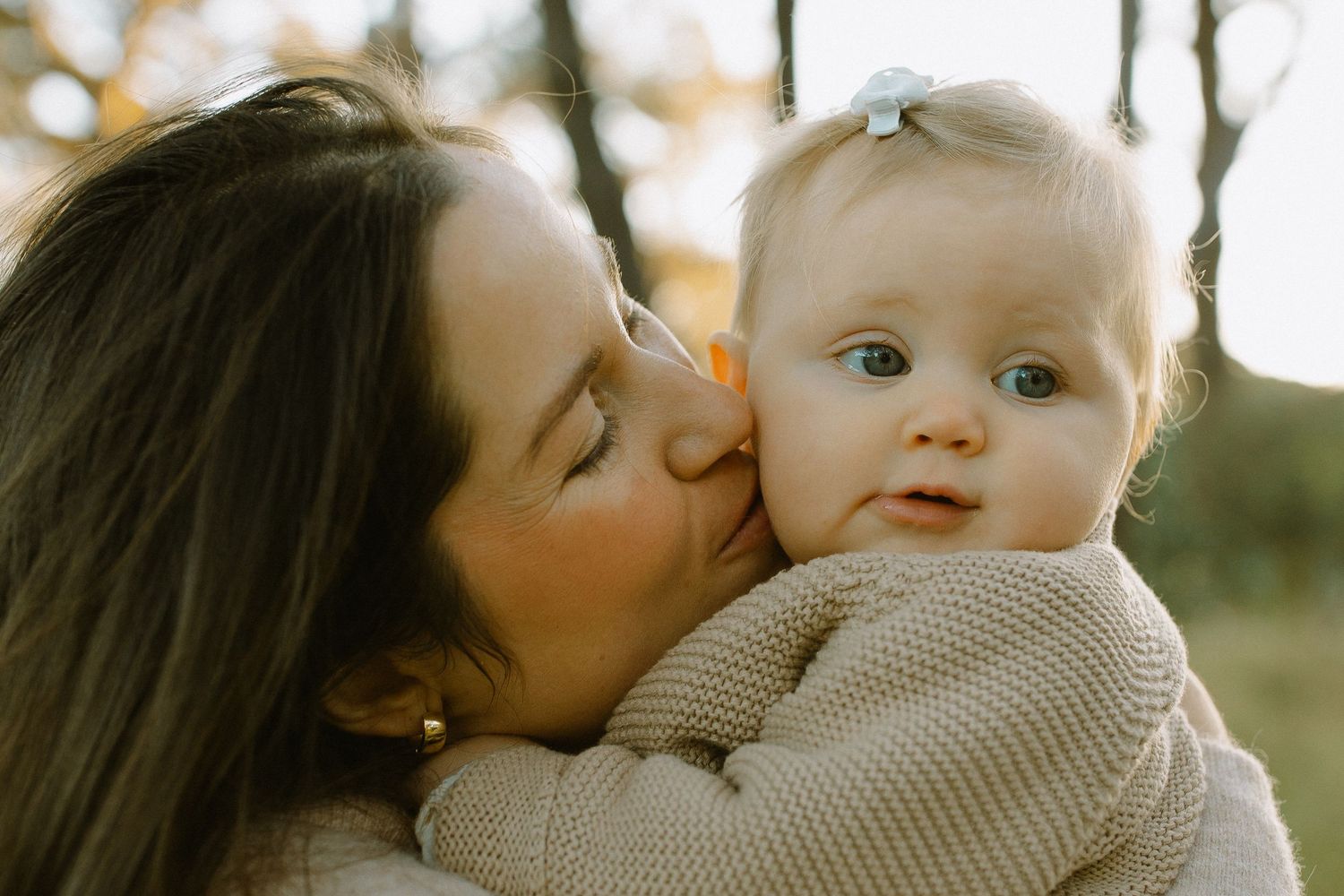 Woman with long dark hair kisses baby in cozy sweater during outdoor photoshoot.
