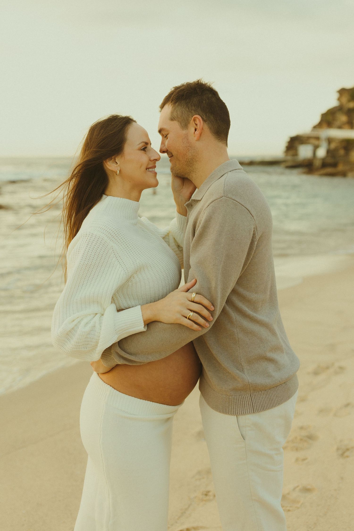 Romantic maternity beach photo of couple in matching cream sweaters embracing against ocean backdrop.