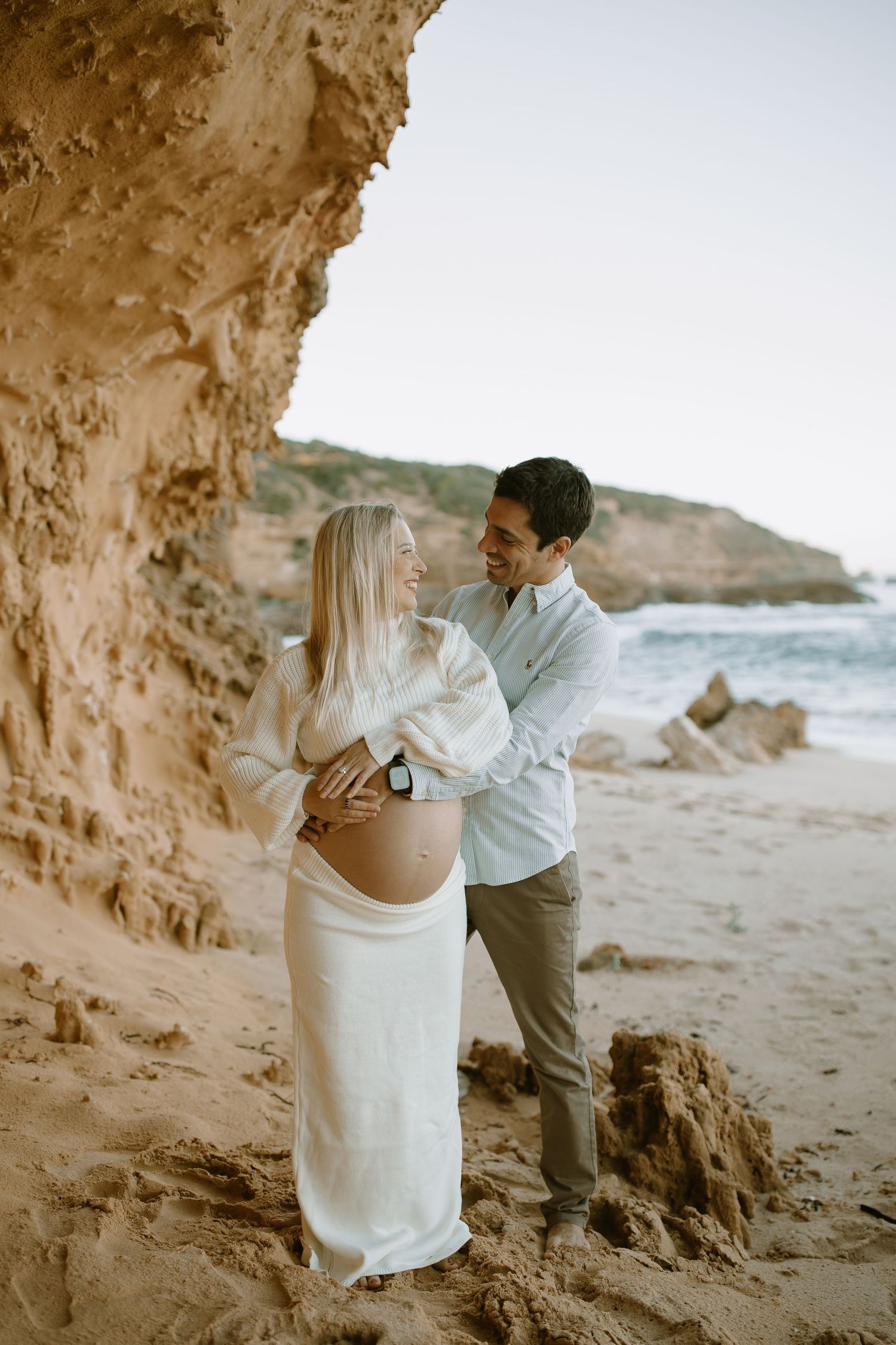 A romantic maternity photoshoot shows a couple embracing under a rocky cave formation at a scenic beach.