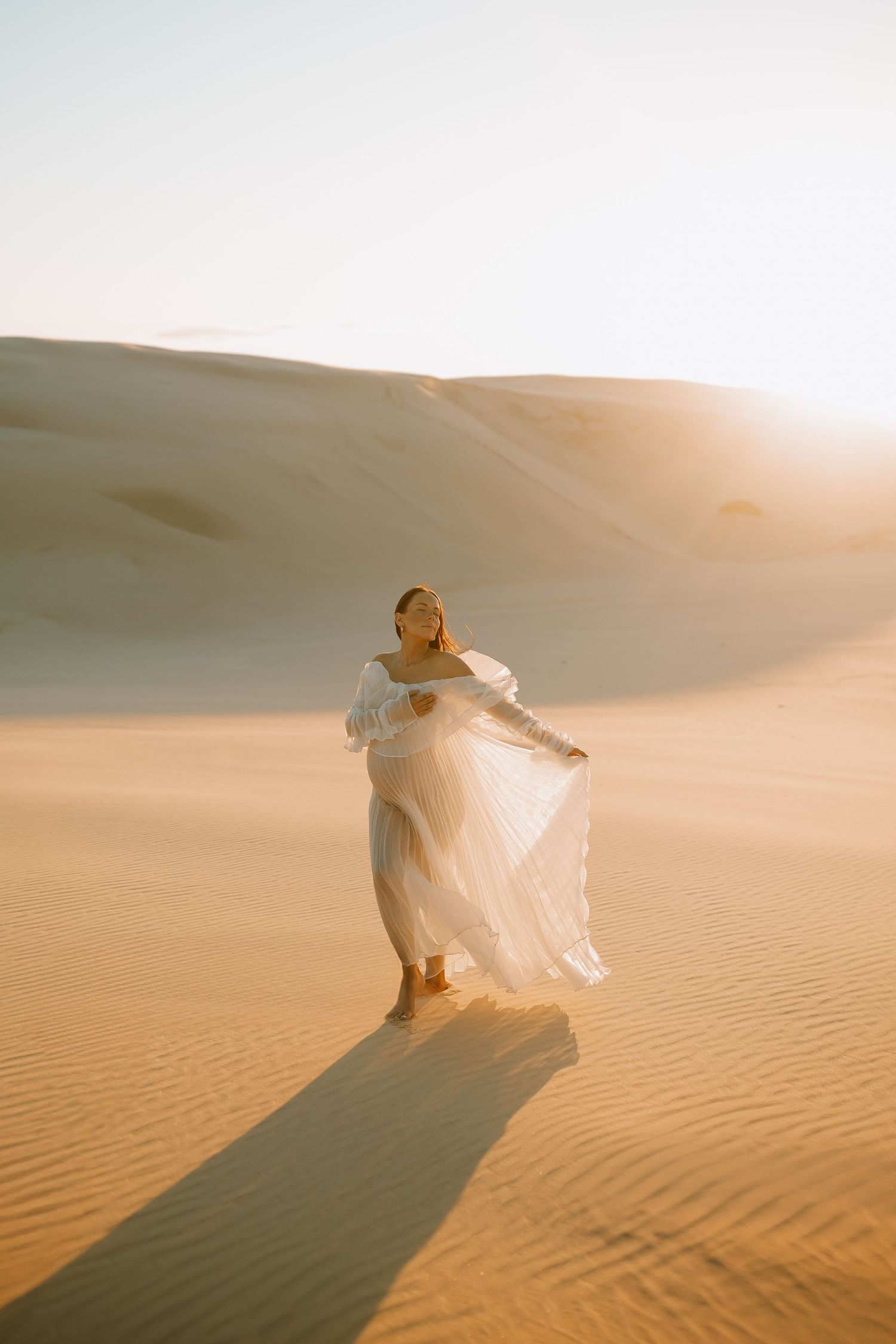 Figure in flowing white dress dancing on sand dunes at sunset casting long shadow in desert landscape.