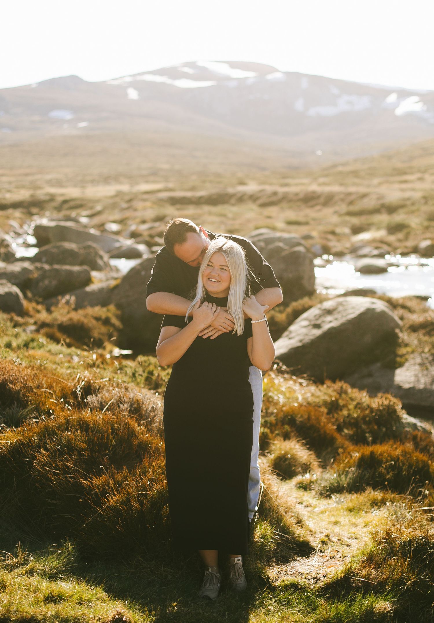 A couple embrace in golden sunlight on a rocky mountain landscape with patches of snow visible in the background.