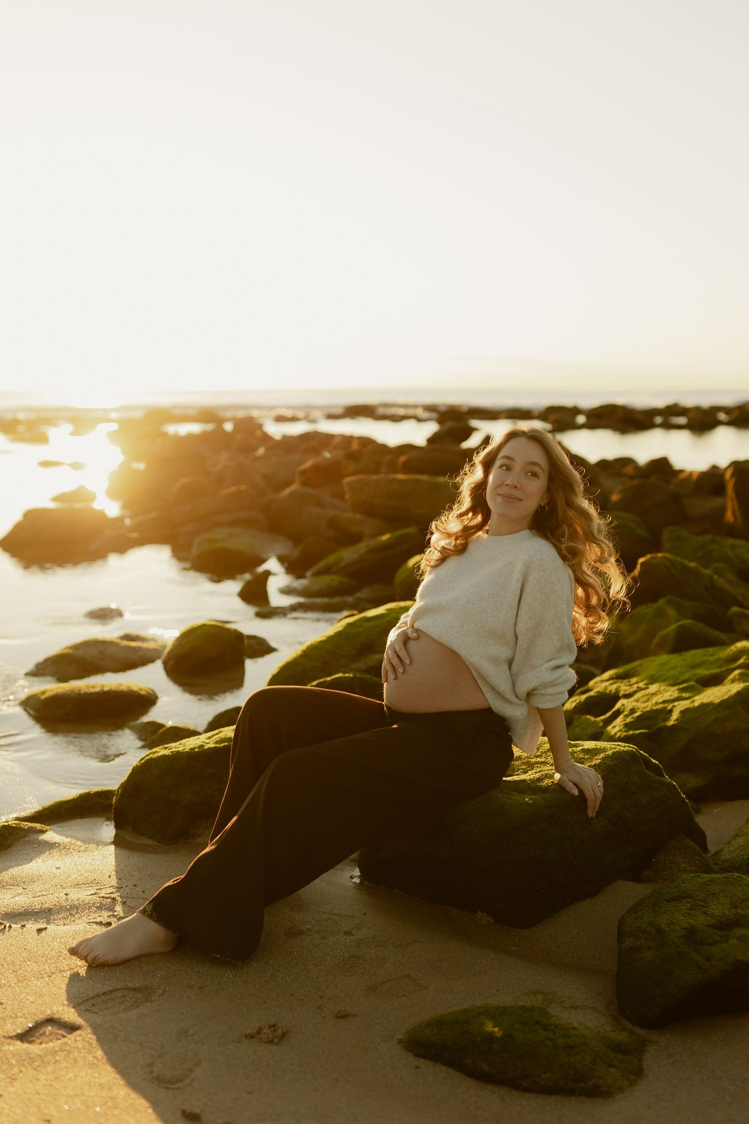 Pregnant woman sitting on moss-covered rocks at golden hour beach sunset, wearing cream sweater and black pants.