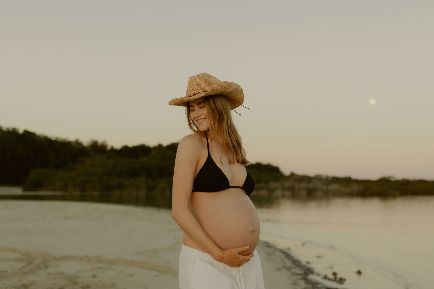 Smiling pregnant woman in straw cowboy hat holds baby bump at sunset beach.