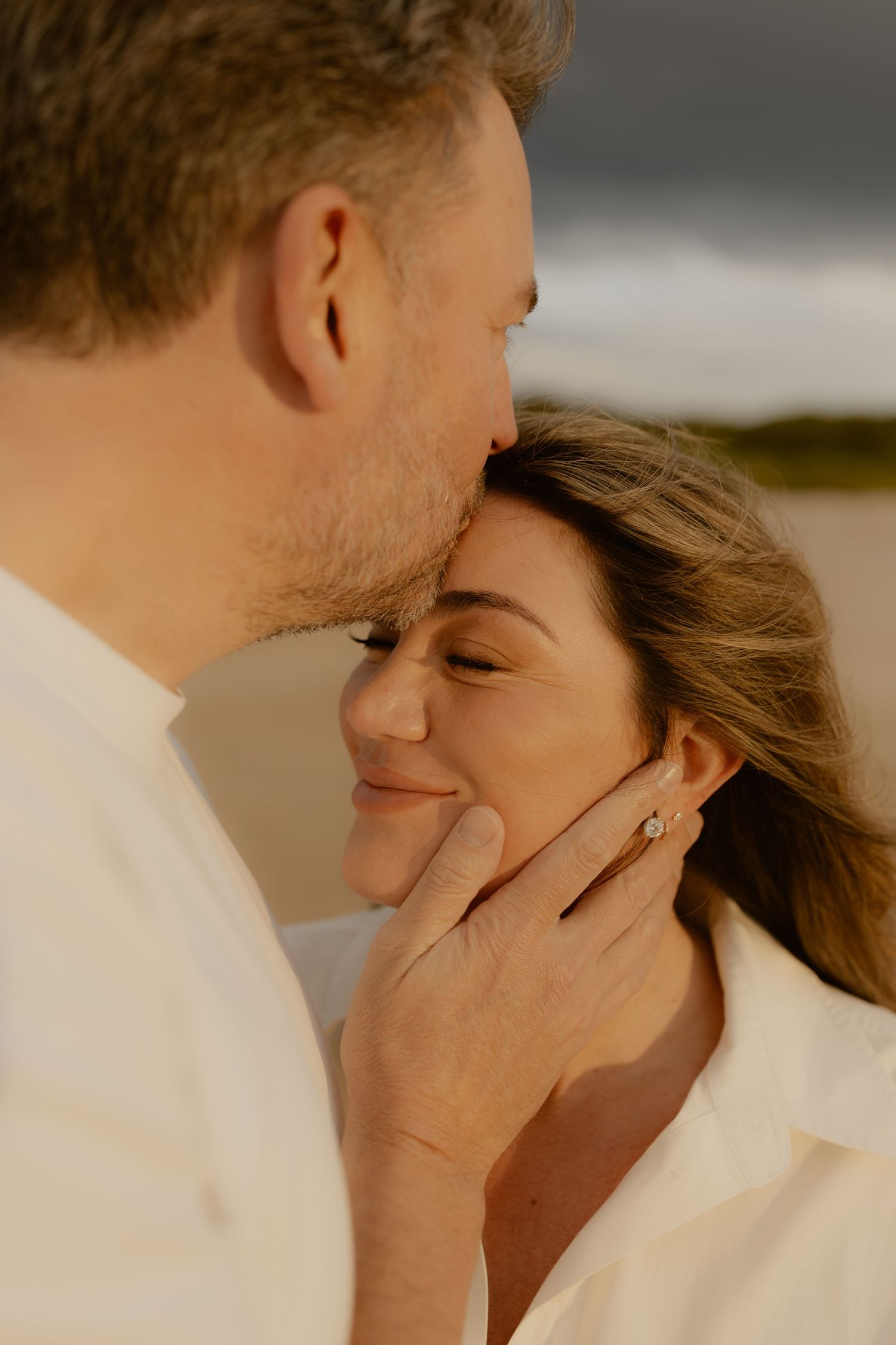 A couple shares a tender moment on a beach, man kissing woman's temple as she smiles, showcasing an engagement ring.