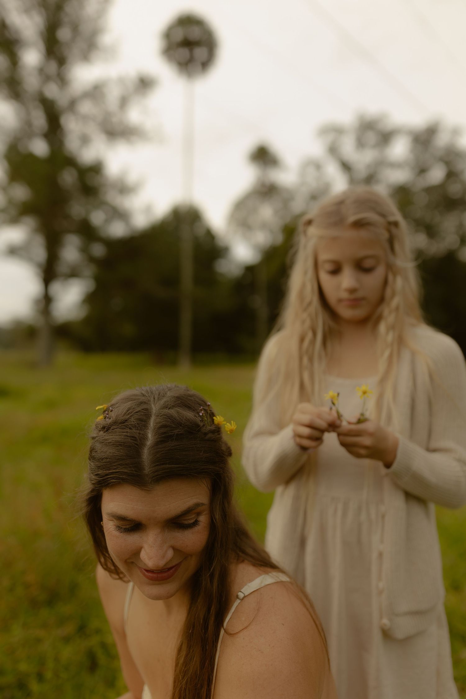 Woman with floral braids and blonde girl holding yellow wildflowers stand in a green pastoral field.