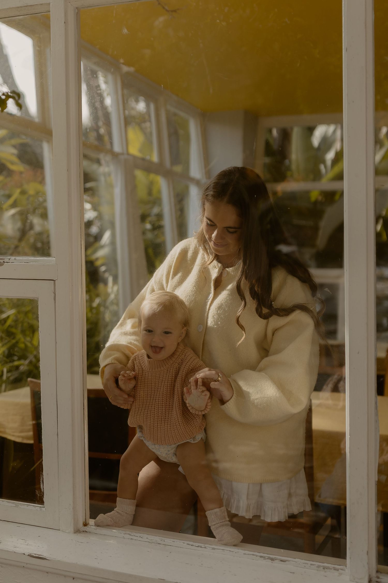 A mother in a cream sweater helps her baby take first steps by a sunlit window surrounded by lush greenery.