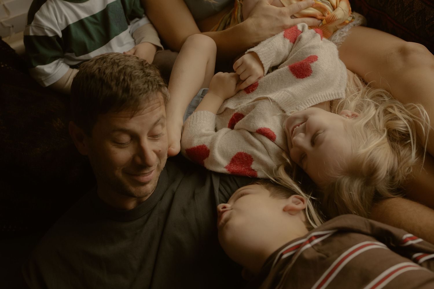 A man and two children lying together on a bed, smiling and relaxing in a cozy, warmly lit room.