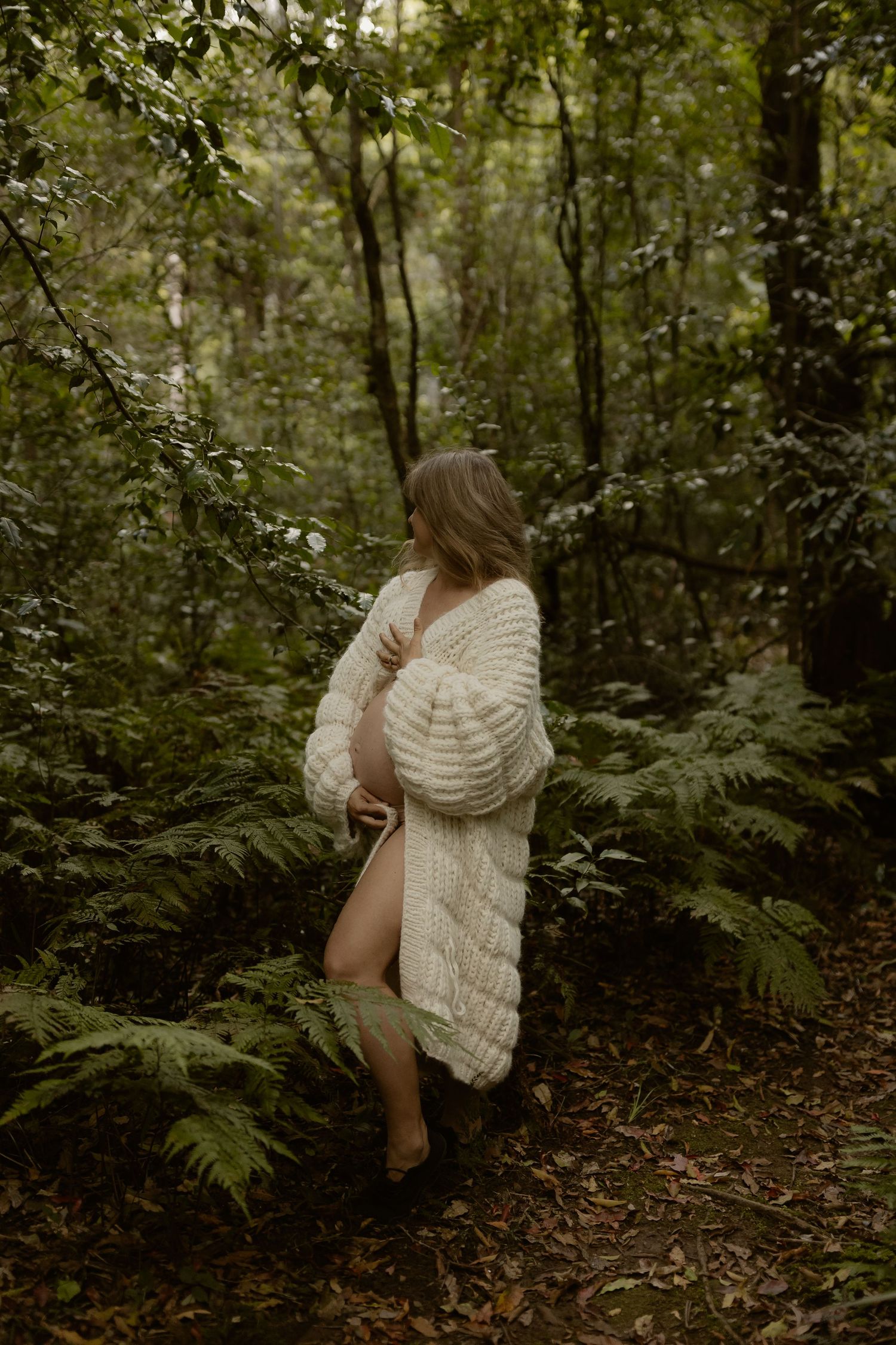 Woman in chunky knit cream cardigan standing among lush green ferns in a forest setting.