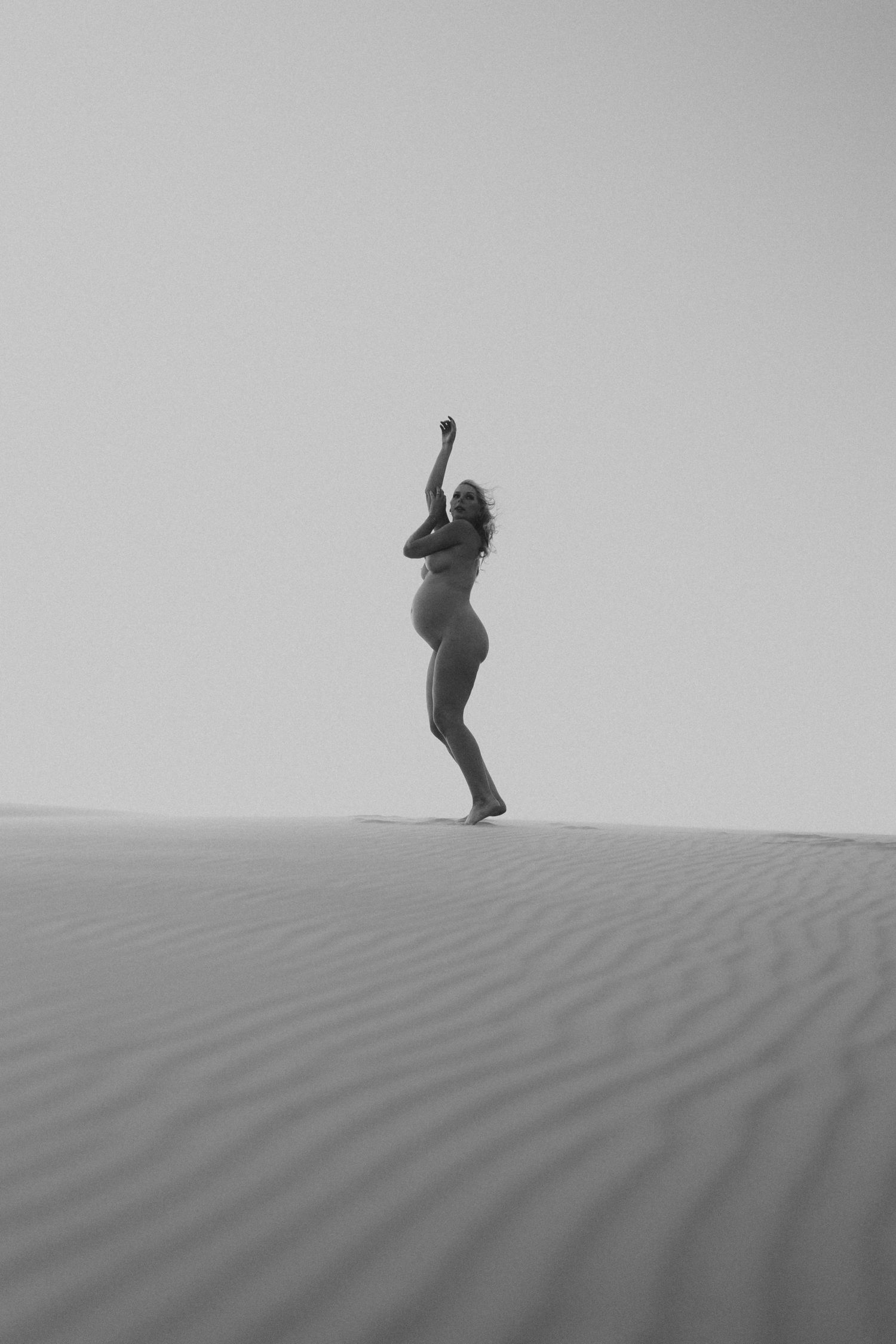 Black and white photo of a pregnant woman posing gracefully on sand dunes in a desert landscape.