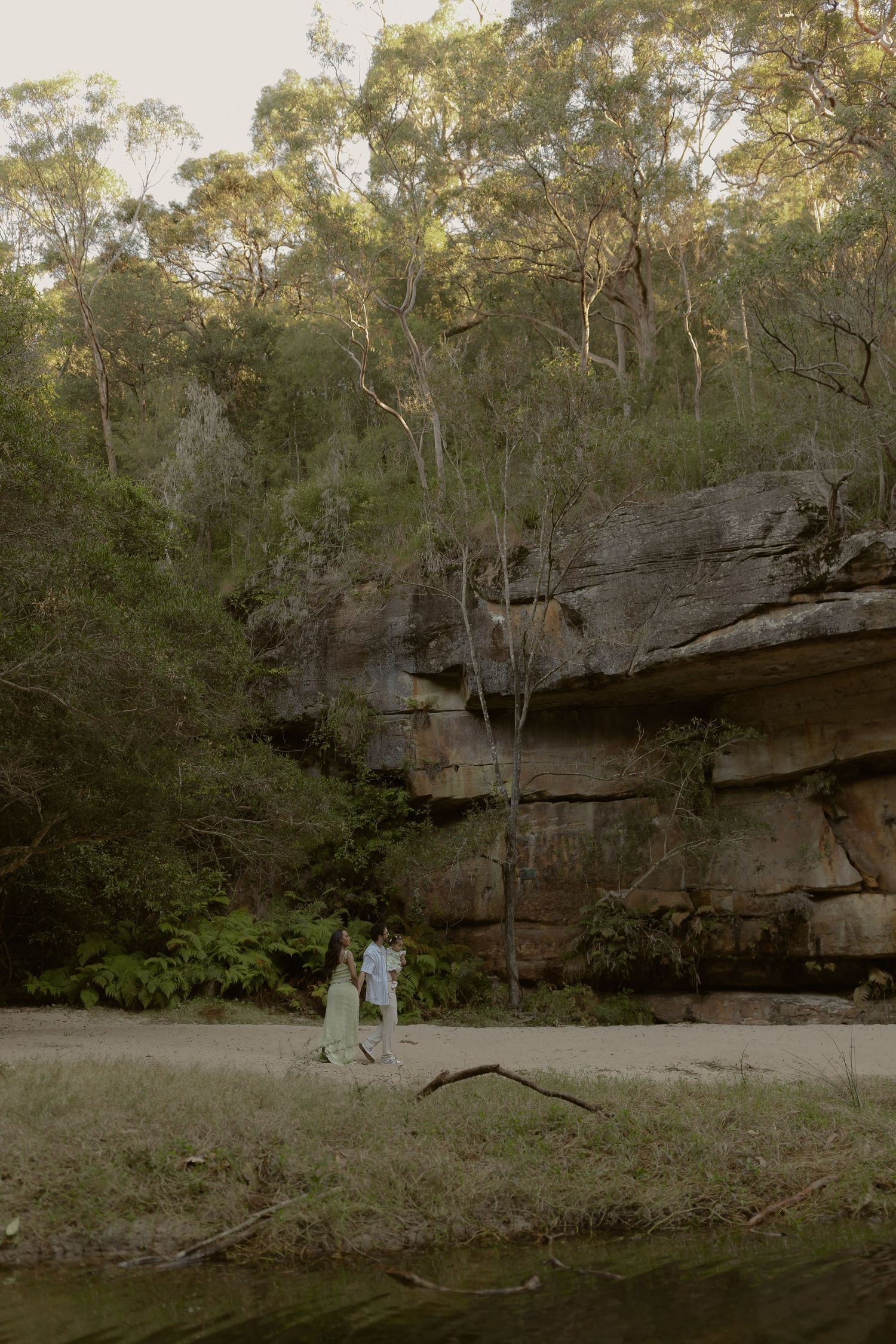 Couple holding hands near sandstone cliffs surrounded by lush green trees beside a tranquil river.