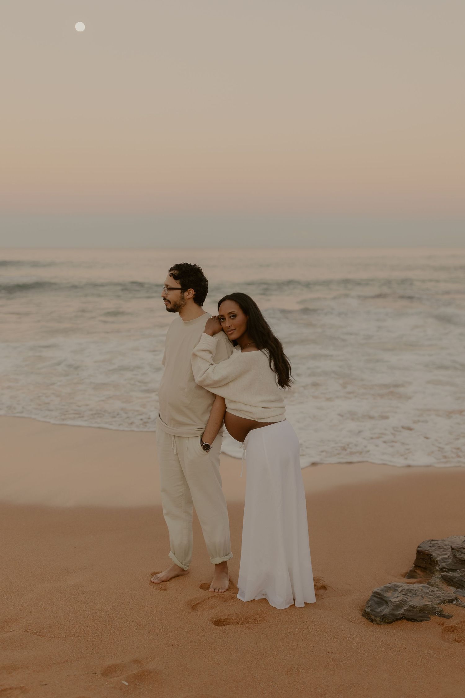 Pregnant couple in neutral tones standing together on sandy beach at dusk with moon above.
