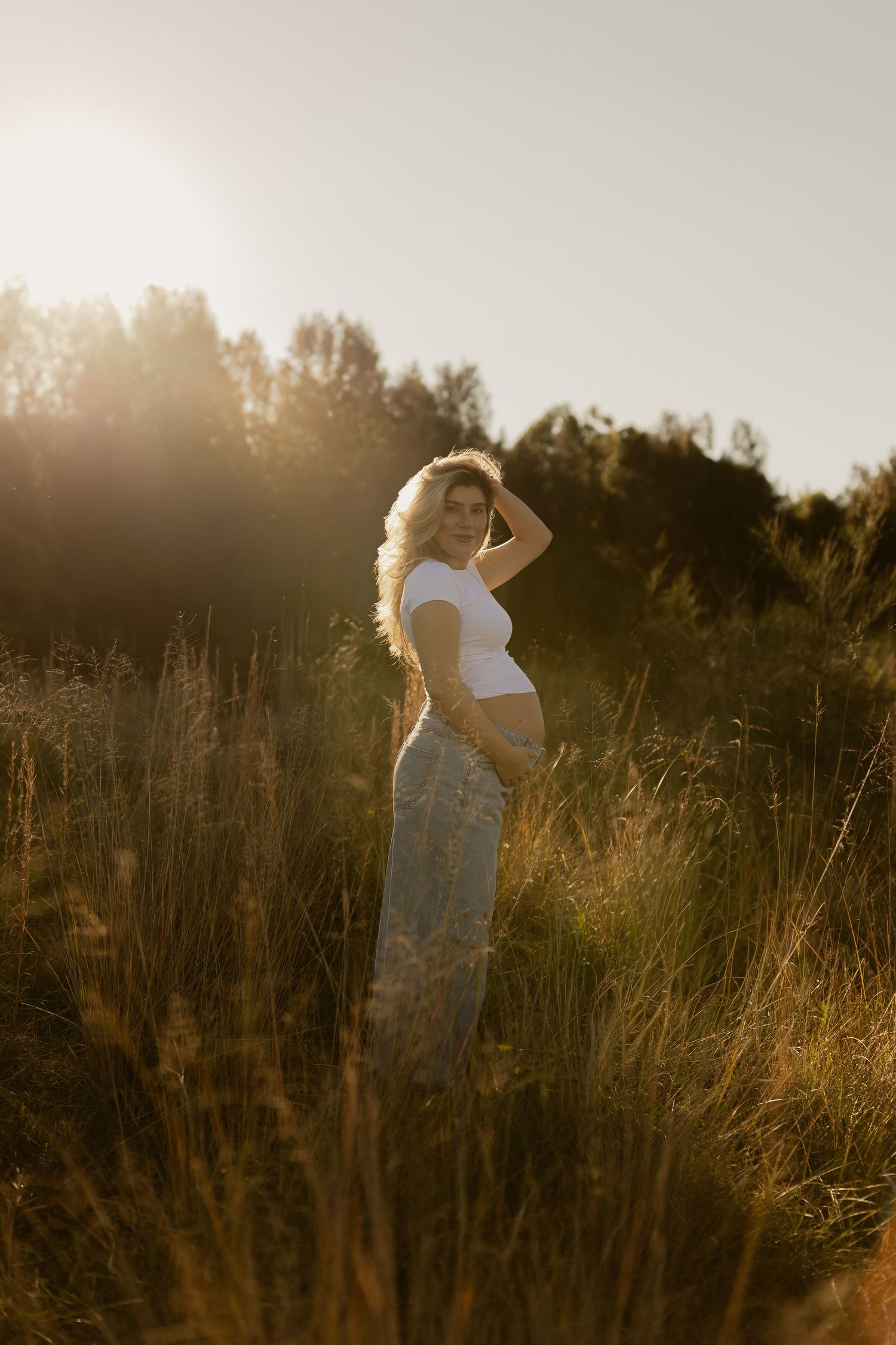 Pregnant woman in white top and floral pants standing in golden sunlit meadow with tall grass and trees at sunset.