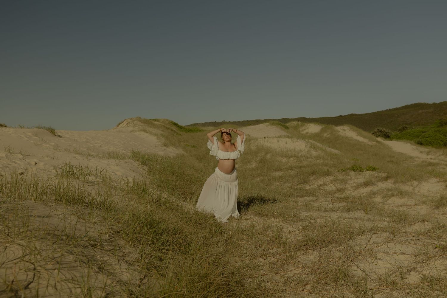 Woman in white dress standing on sandy dunes with green grass under a clear sky.