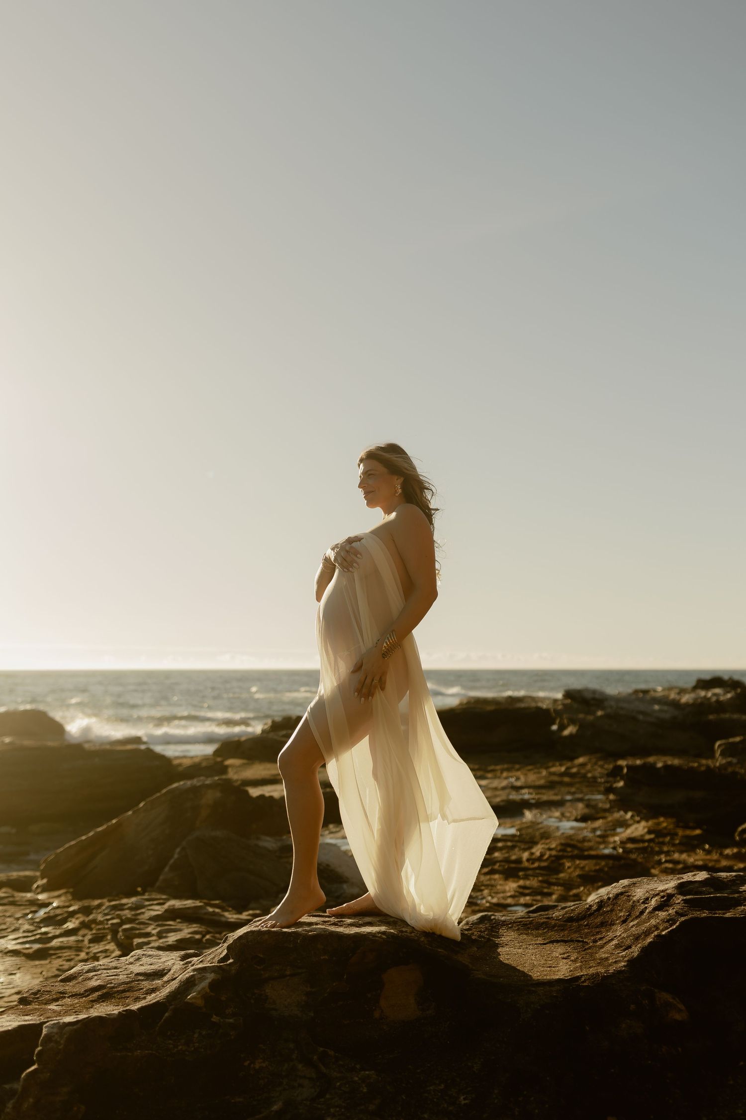 A woman draped in sheer white fabric stands on coastal rocks at golden hour with ocean waves in the background.