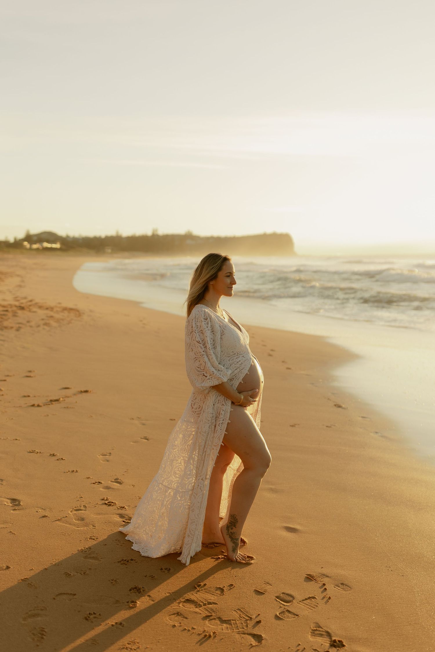 Pregnant woman in white lace gown cradling baby bump on golden sandy beach at sunset.