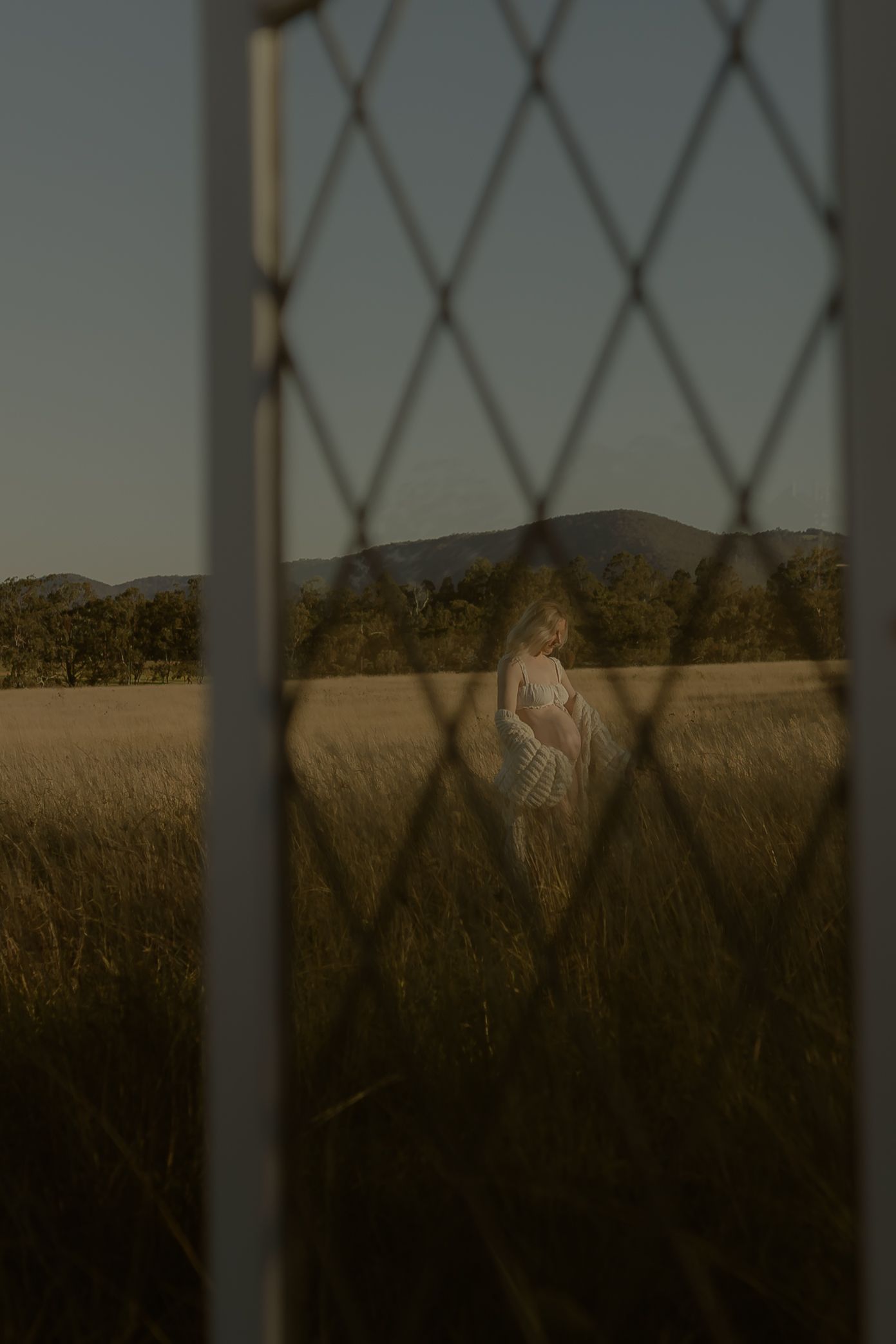 A woman stands in a golden field viewed through a diamond-pattern wire fence at dusk, with hills and trees behind her.