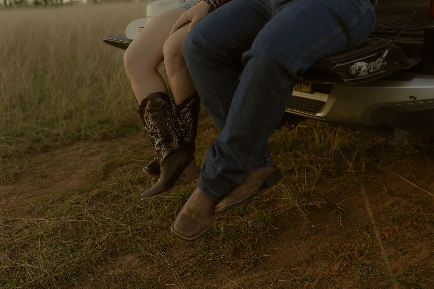 Two people sitting on a truck tailgate wearing cowboy boots and jeans in a rustic field at golden hour.